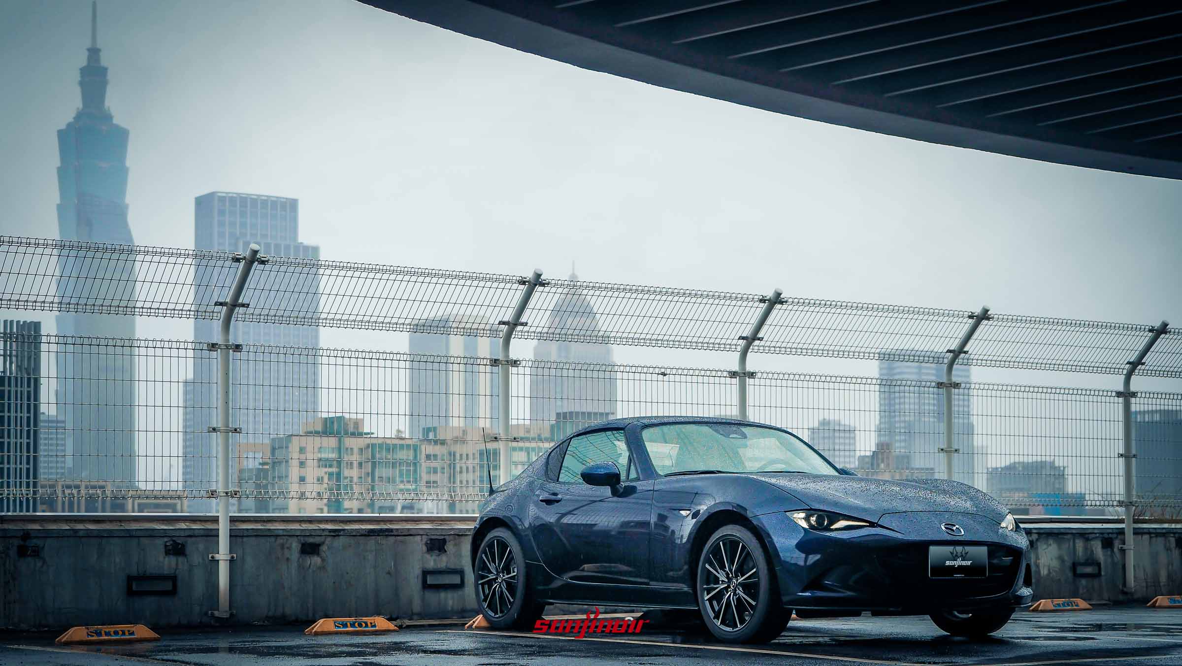 一輛深藍色的 Mazda MX-5 RF 停在雨中的頂樓停車場，背景為霧霾中若隱若現的台北101大樓。