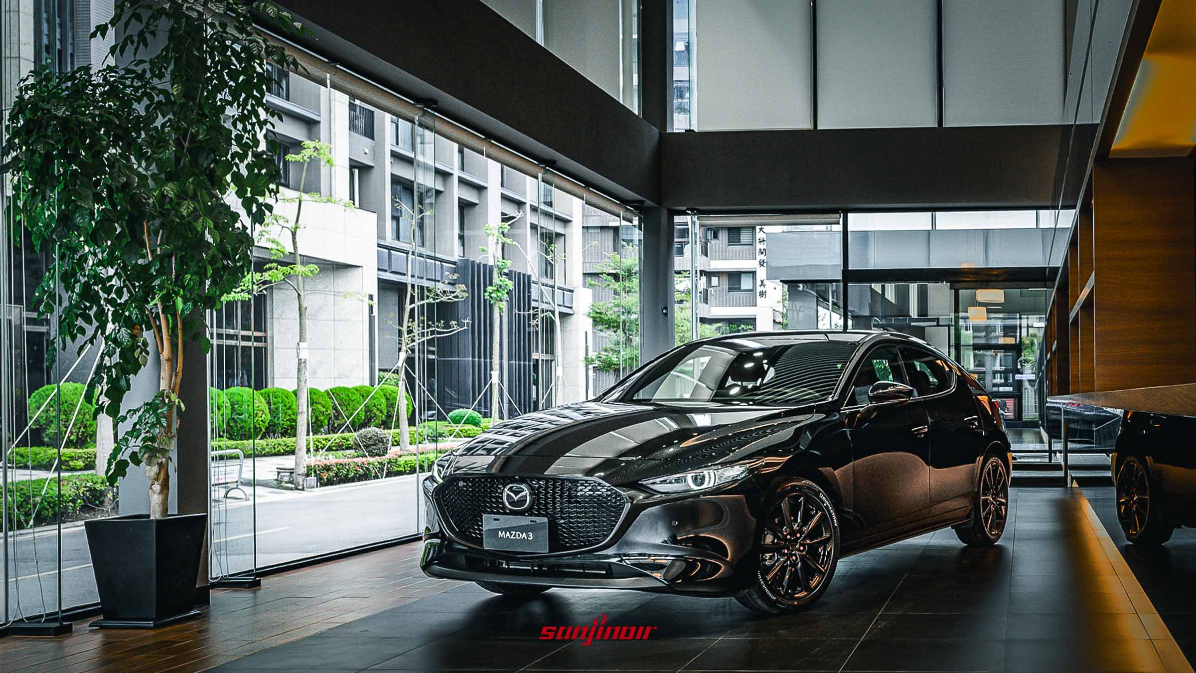 黑色 MAZDA3 HOMURA 五門掀背車型，配備黑化水箱護罩與爍黑鋁合金輪圈，停放於明亮的 MAZDA 展示中心內。