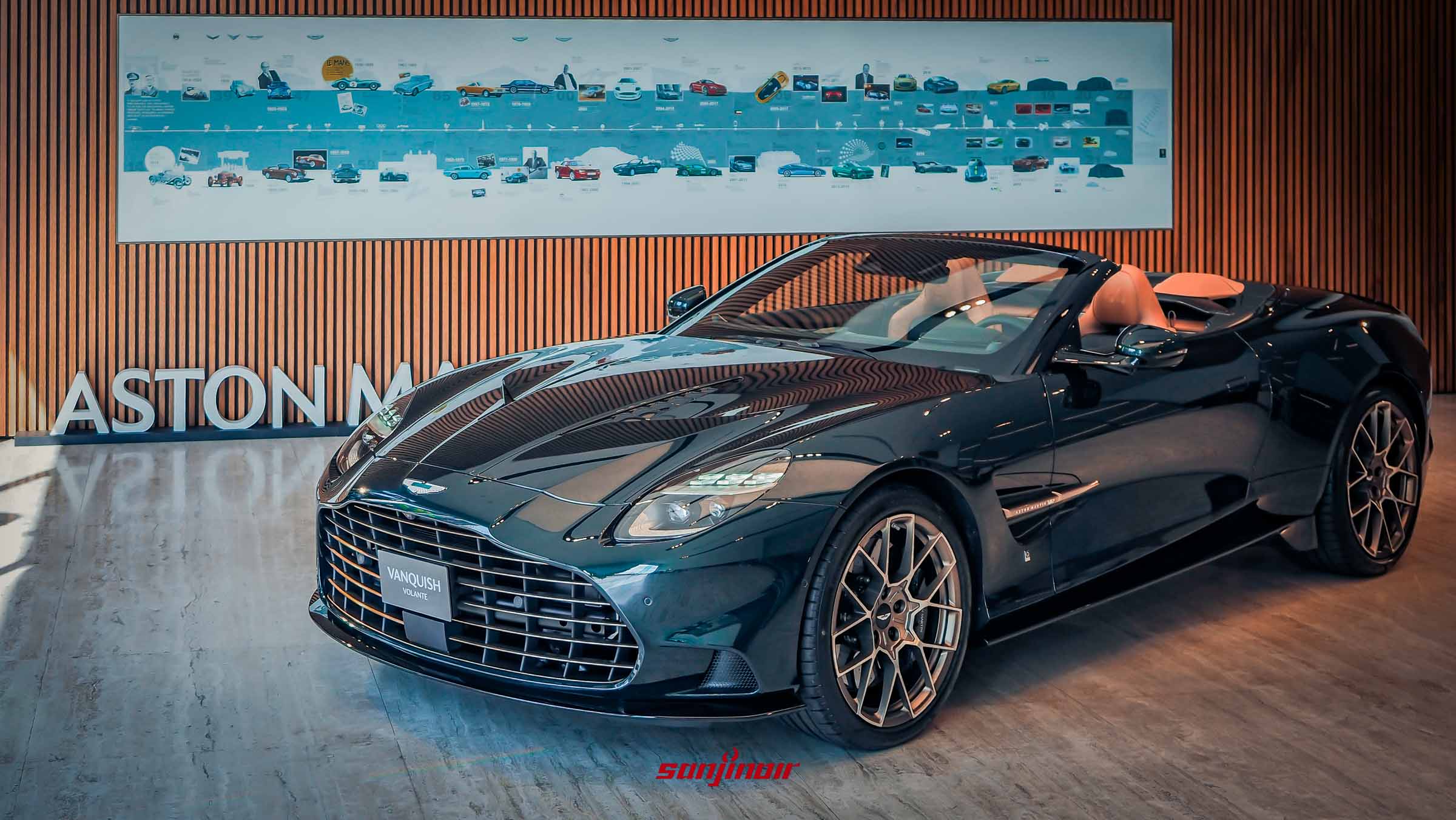 一輛深綠色（Pentland Green）的 Aston Martin Vanquish Volante 60th Anniversary Edition 敞篷車，軟頂收起，停在有木質格柵背景和展示 Aston Martin 歷史車型看板的展場。車輛為 45 度角前視，可見馬鞍棕色內裝和頭枕上的 60 週年刺繡。