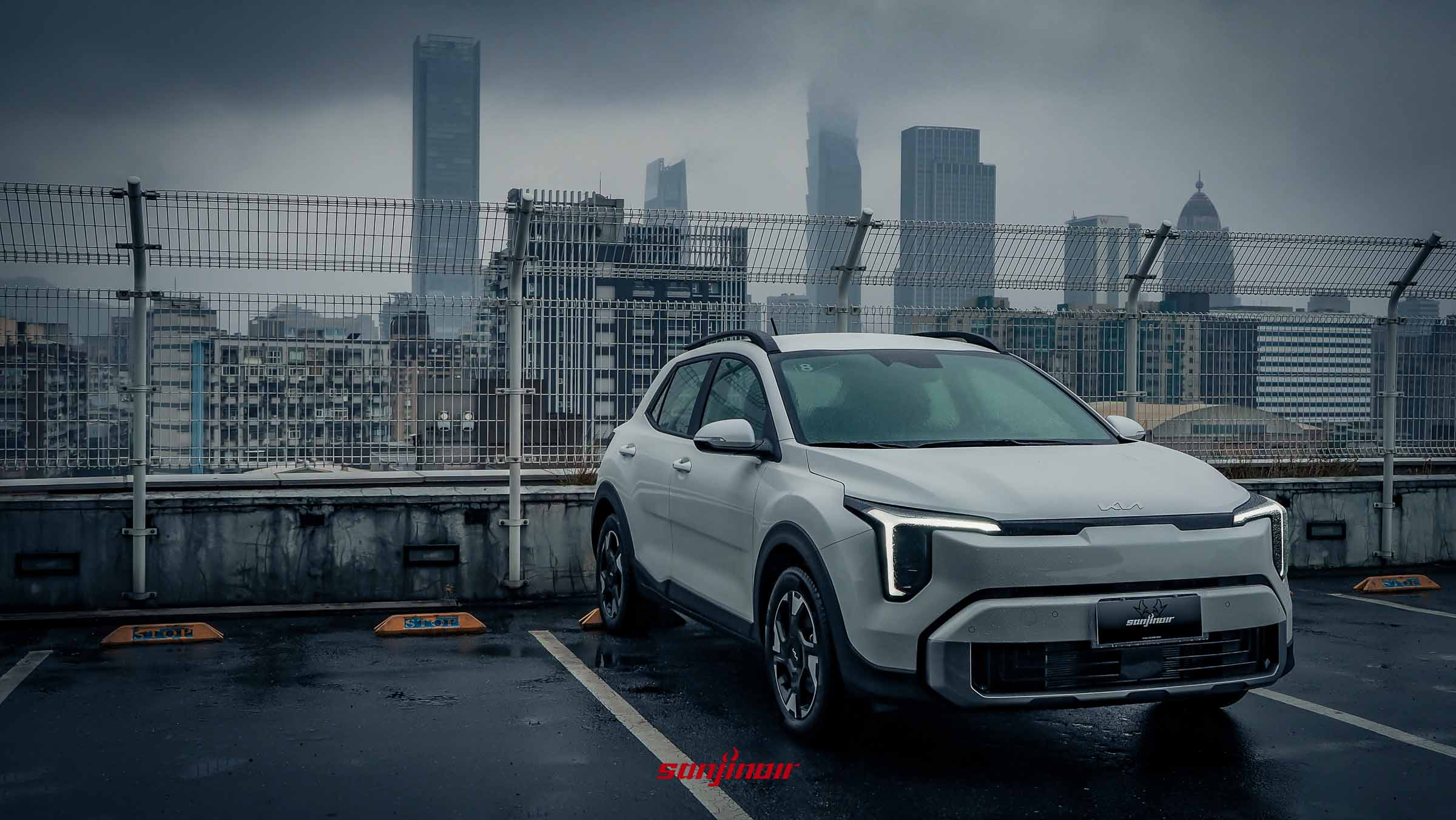 停放在頂樓停車場的白色2026 Kia Stonic跨界休旅車前方斜側面照，背景為陰雨天的城市高樓。