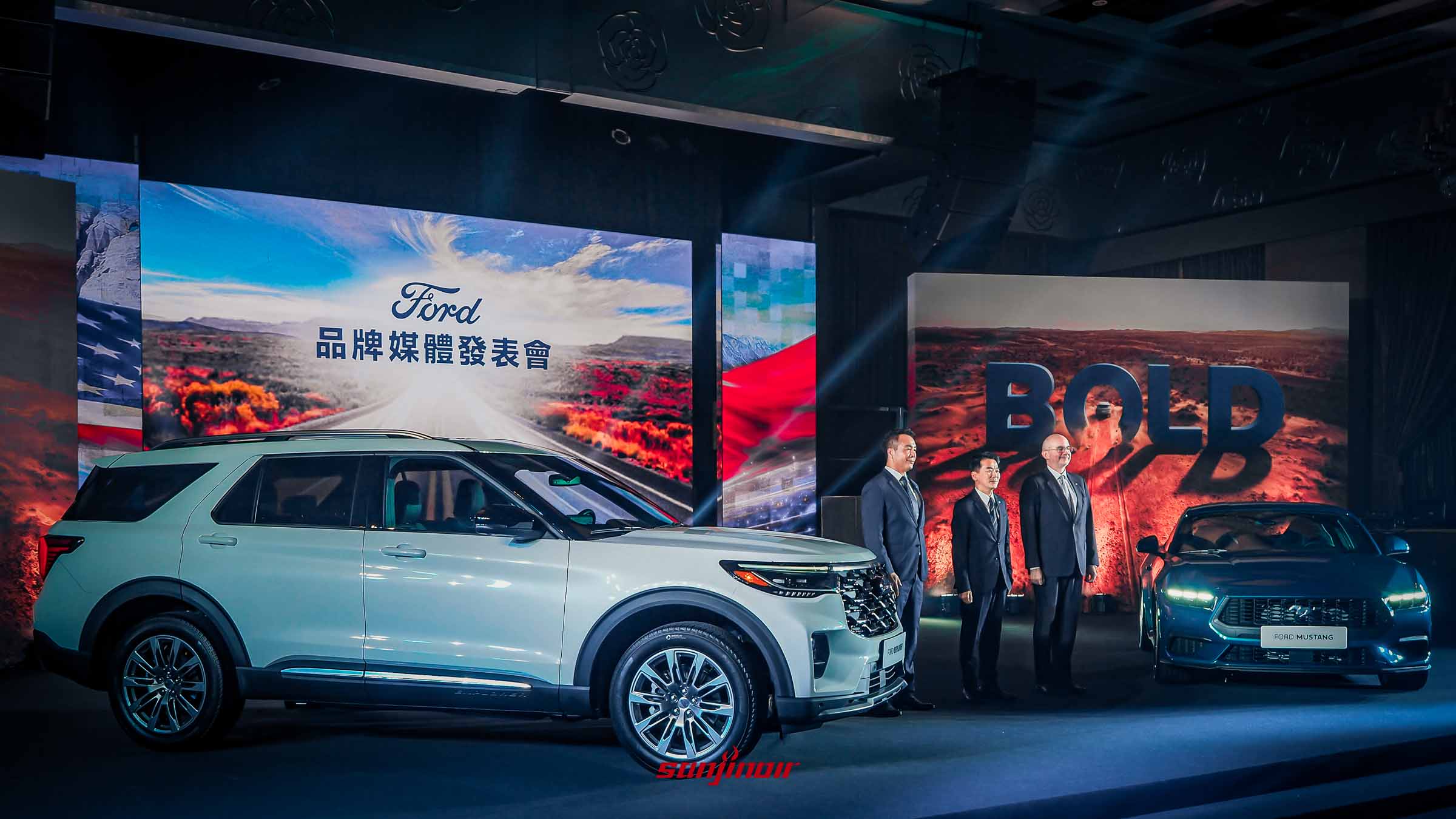 福特「GO BOLD GO FORD」品牌媒體發表會現場全景。舞台左側停放一輛白色Ford Explorer，右側停放一輛藍色Ford Mustang。三位西裝革履的男士（AIT和福特代表）站在中央。背景大螢幕顯示「Ford 品牌媒體發表會」和「BOLD」字樣。