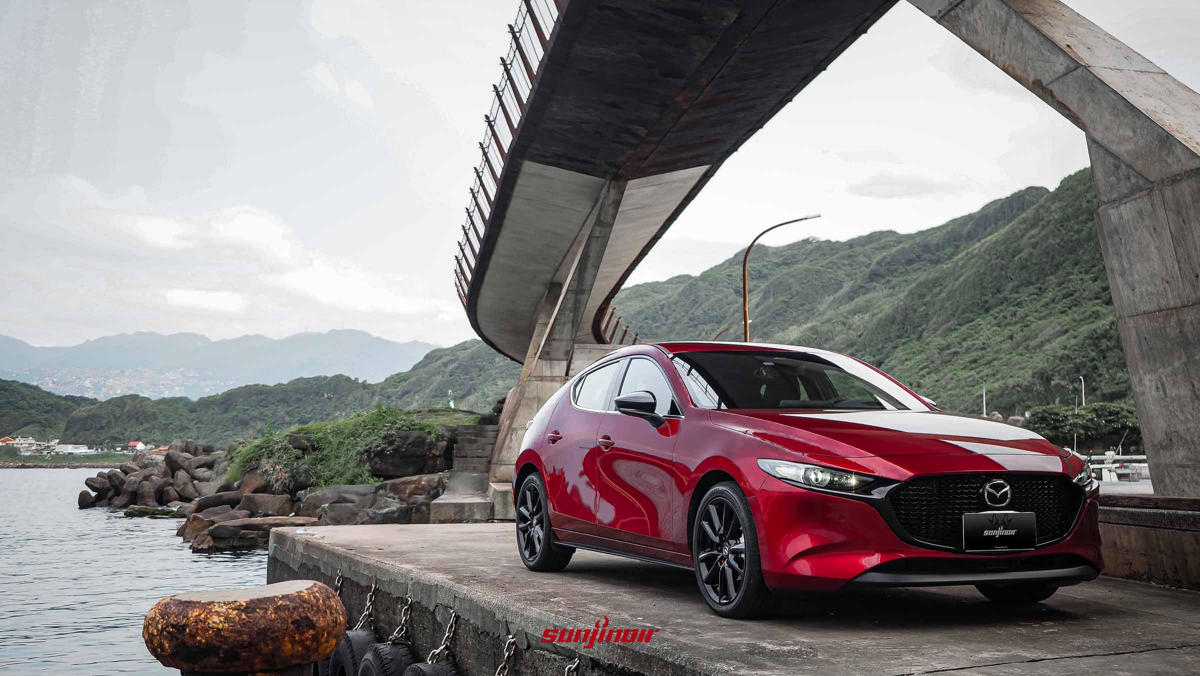 一輛晶艷魂動紅MAZDA3掀背車停在海邊碼頭上，背景是混凝土高架橋和綠山。