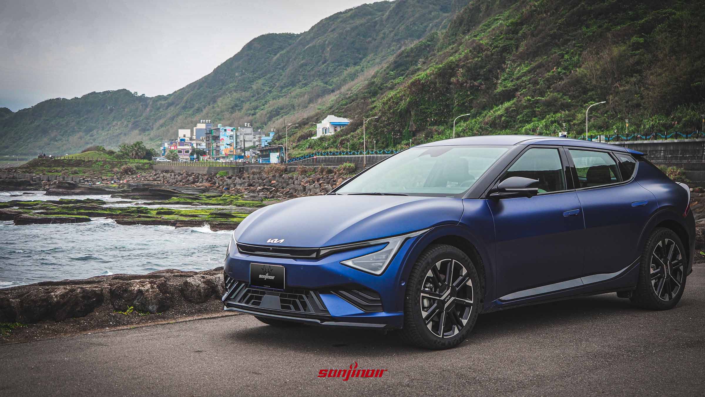 藍色啞光Kia EV6 GT-Line電動車停在海邊道路上，背景是連綿的山脈。