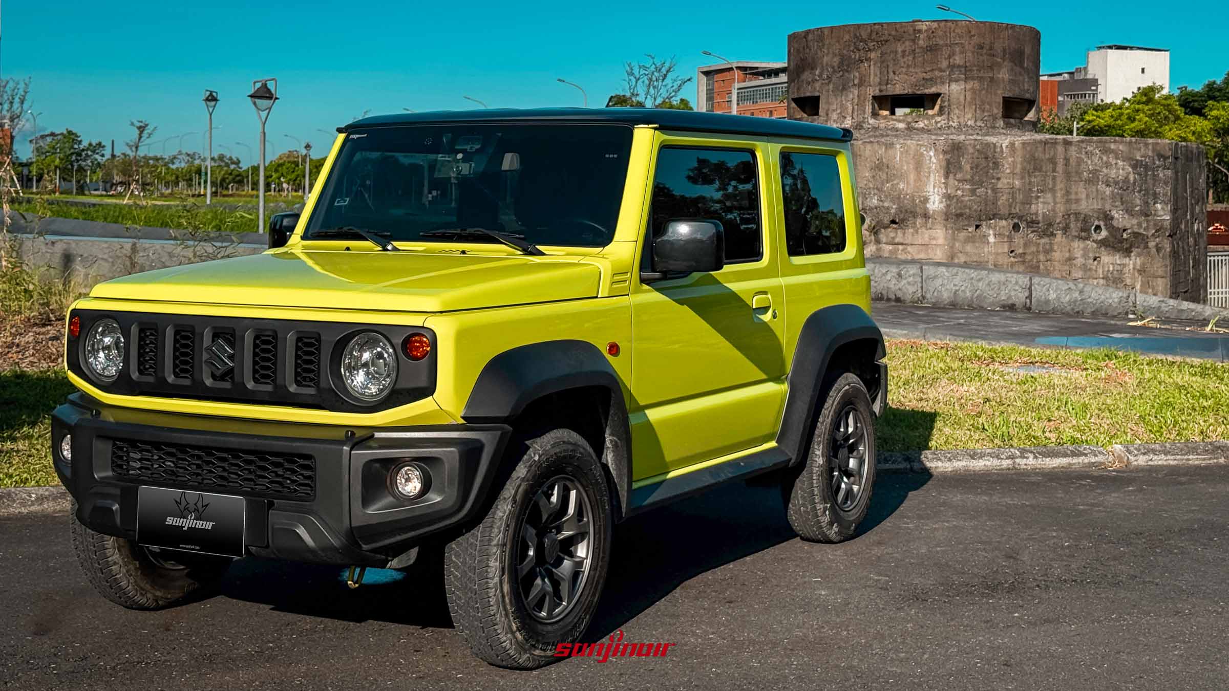 一張戶外拍攝的亮檸檬綠 SUZUKI Jimny JB74（配黑頂）前右側高角度照片，停在柏油路上。背景為陽光明媚的綠地公園、路燈和一座混凝土碉堡結構。