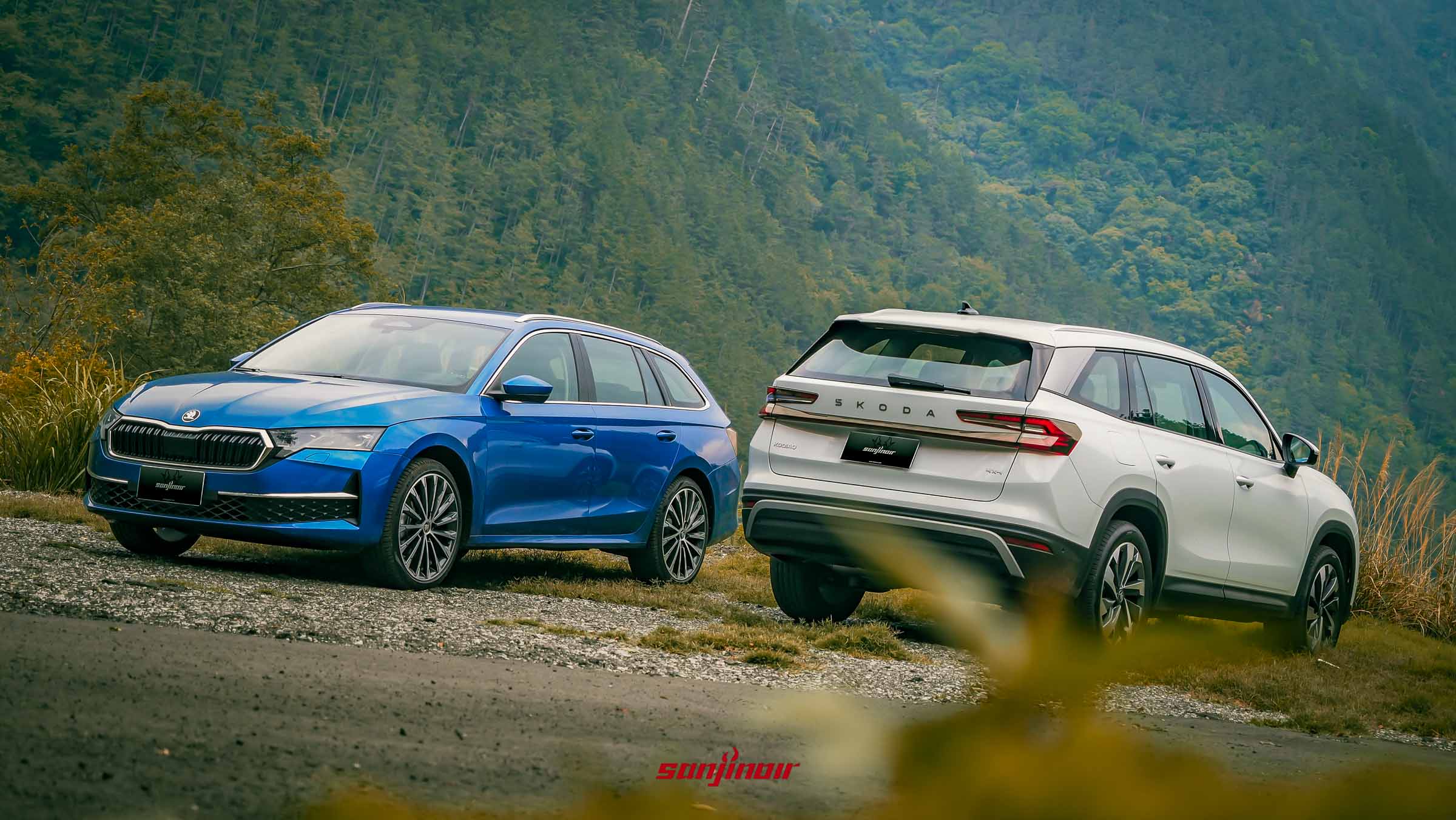 Škoda Octavia Combi 2.0 TSI 4×4（藍色）和 Škoda Kodiaq 2.0 TSI 4×4（白色）並排停在礫石路面上的全景照片，背景是茂密的松樹林和陡峭的山脈。Kodiaq 顯示車尾。