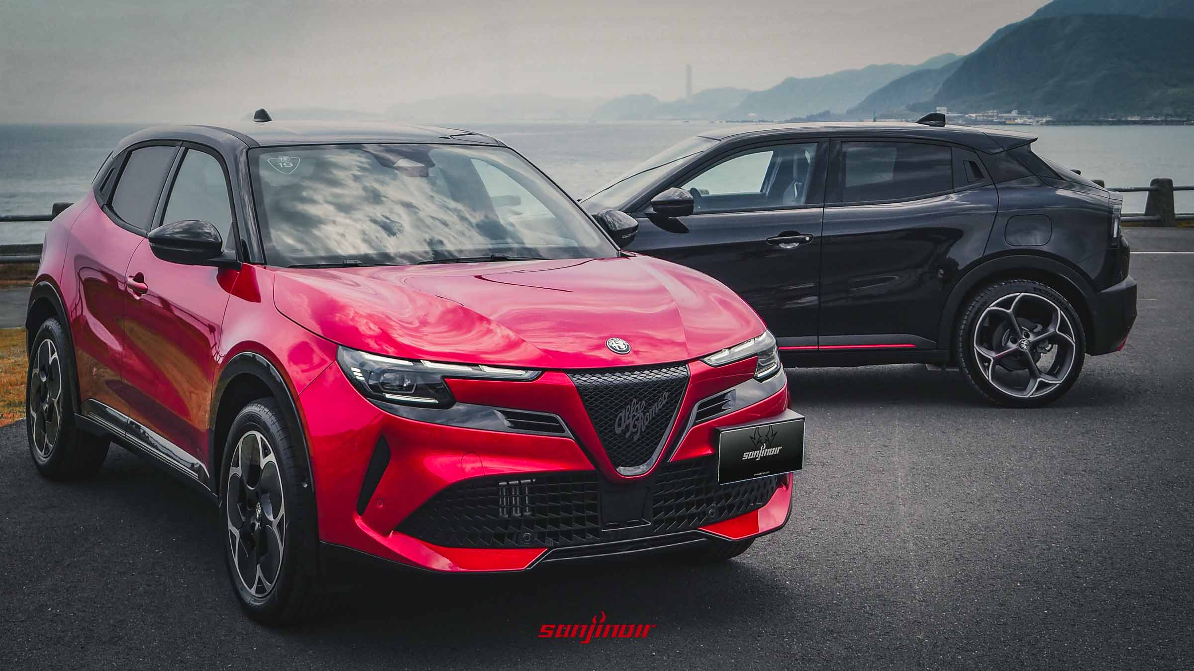 兩輛Alfa Romeo Junior停在海岸邊，前方為紅色車型，後方為黑色車型，展示車身輪廓與流暢線條。