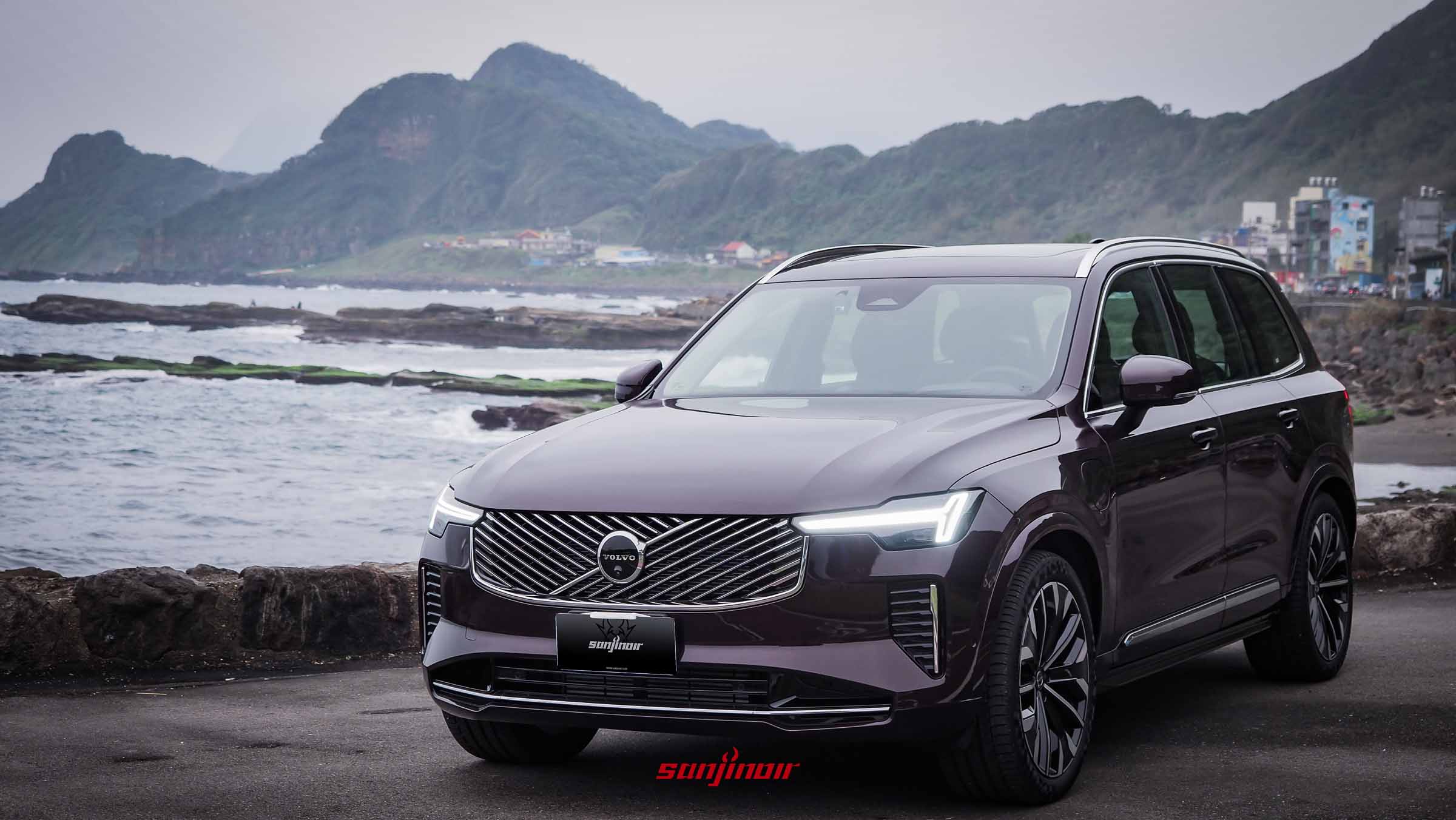 一輛深紫黑色的Volvo XC90 T8 Ultra大型豪華休旅車停在海岸道路旁，背景為陰天下的山脈與海面。