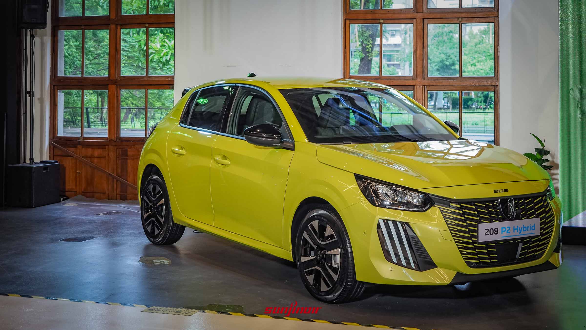 一輛鮮黃色的PEUGEOT 208 P2 Hybrid停放於帶有木窗的室內展演空間，自右前方45度角拍攝，展現俐落車身線條與獨特雙色鋁圈。