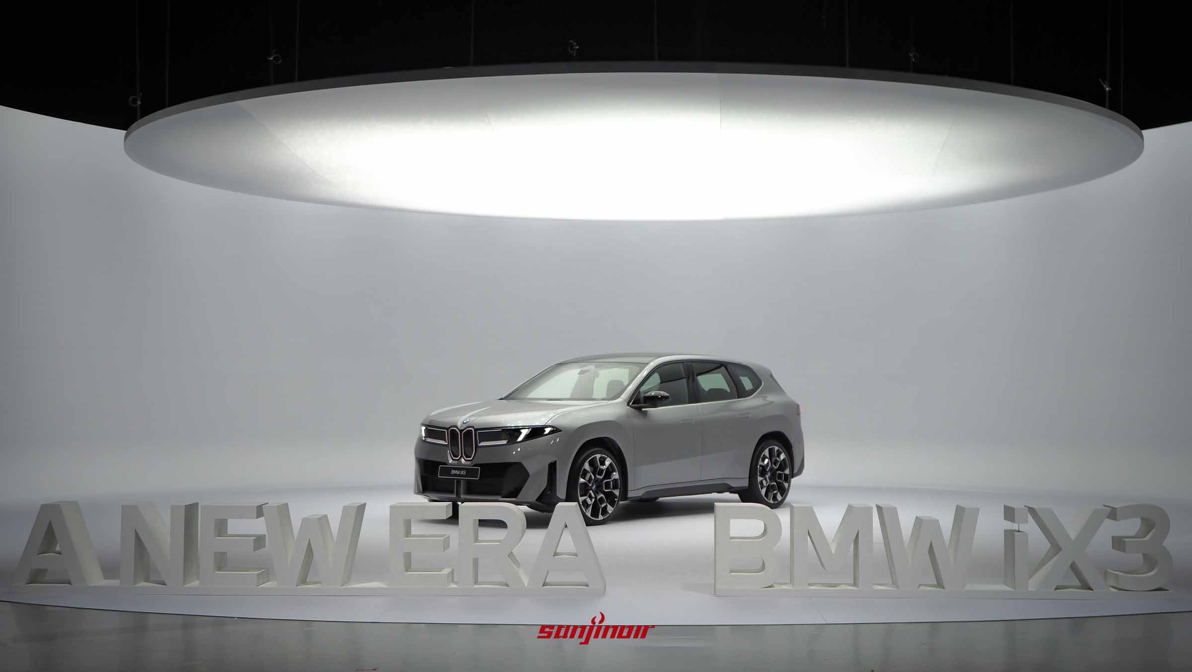 圓形舞台中央停放著一輛銀灰色的BMW iX3純電休旅車，前方擺放立體白色英文字樣。