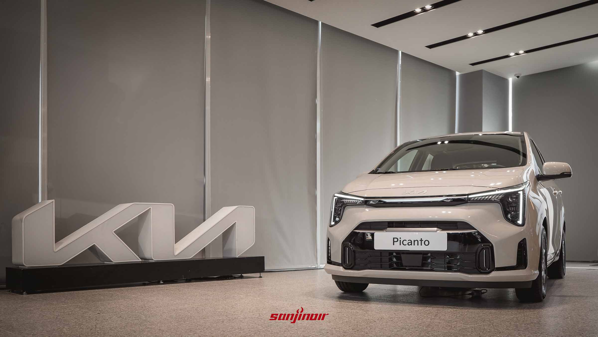 一輛停放在明亮展示中心的Kia Picanto Cappuccino Edition特仕版微型車，車身為牛奶白，旁邊擺放著大型Kia立體廠徽。