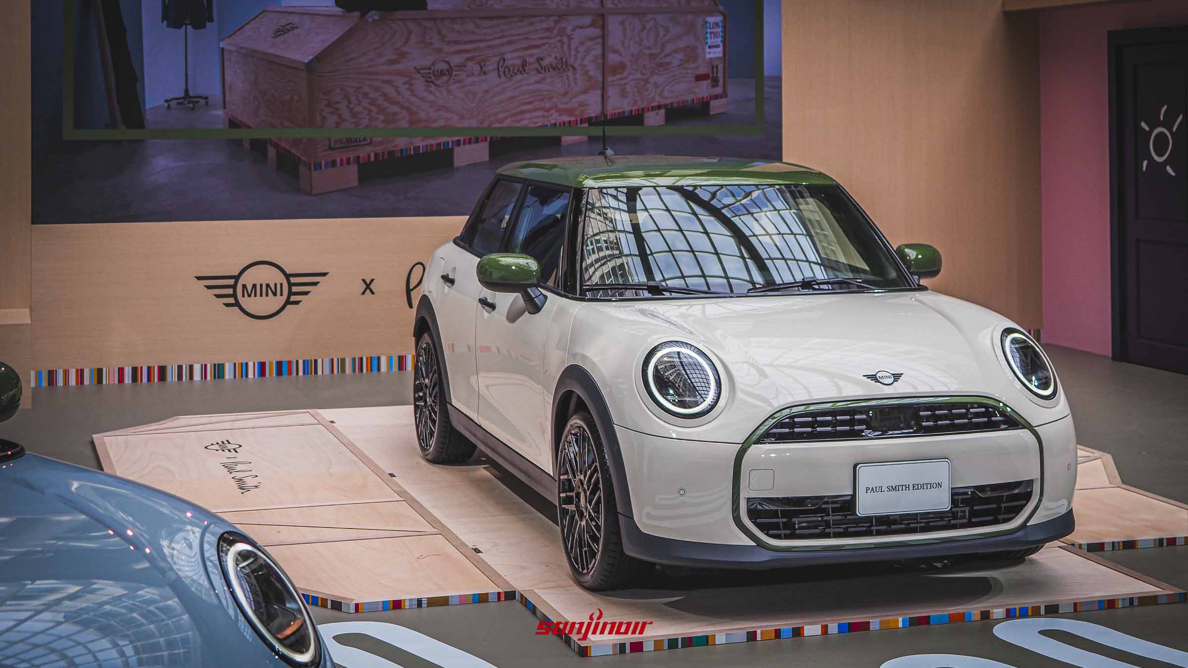 一輛白色的MINI Paul Smith Edition停放在木製展示台上，車頭水箱護罩外框和兩側後視鏡為綠色，背景有MINI x Paul Smith的聯名標誌。