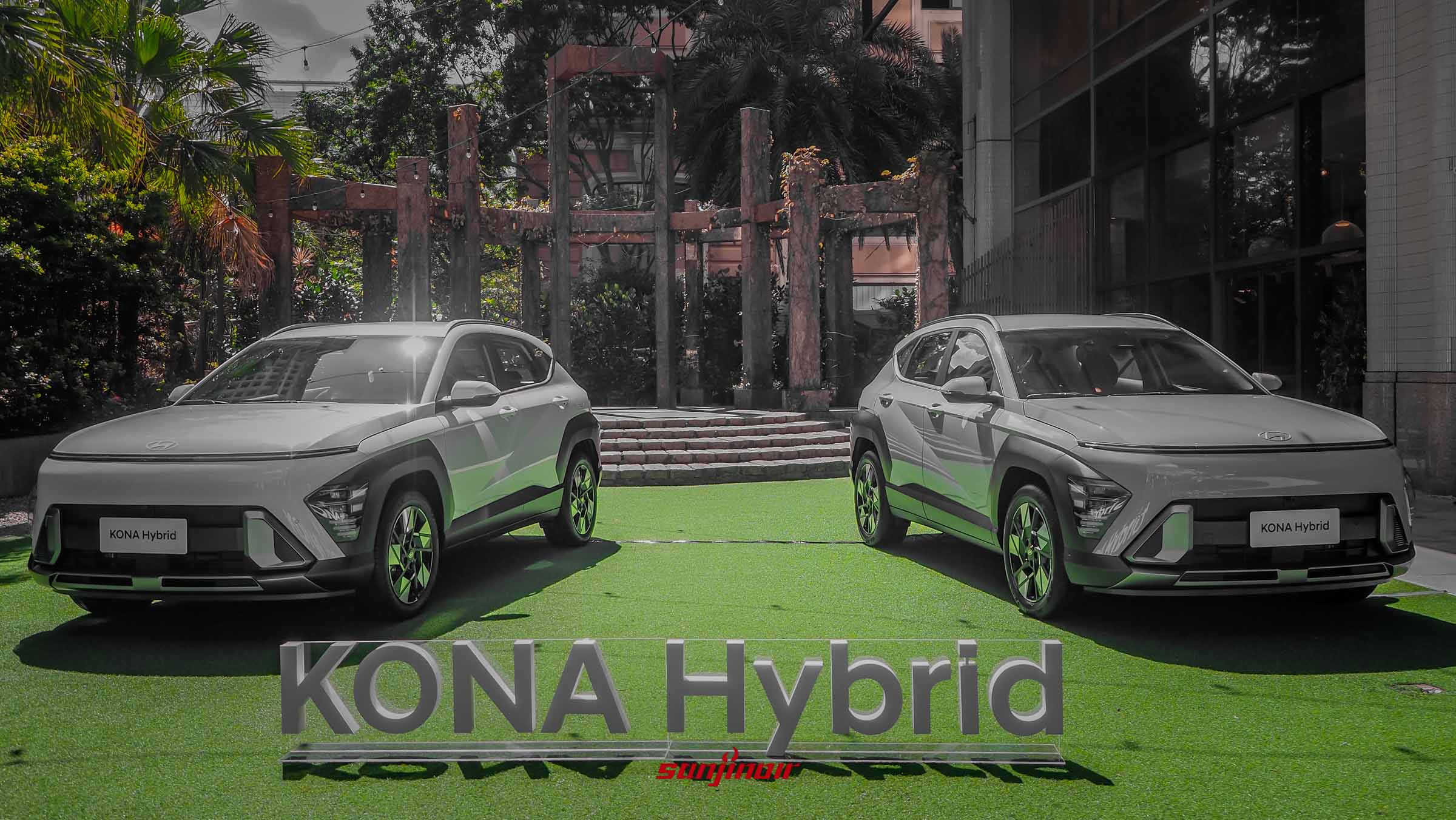 兩輛淺灰色的2026 Hyundai Kona Hybrid並排停放於充滿綠意的戶外草皮上，前方立有透明壓克力材質的「KONA Hybrid」字樣，背景為帶有工業風格的建築與植物。
