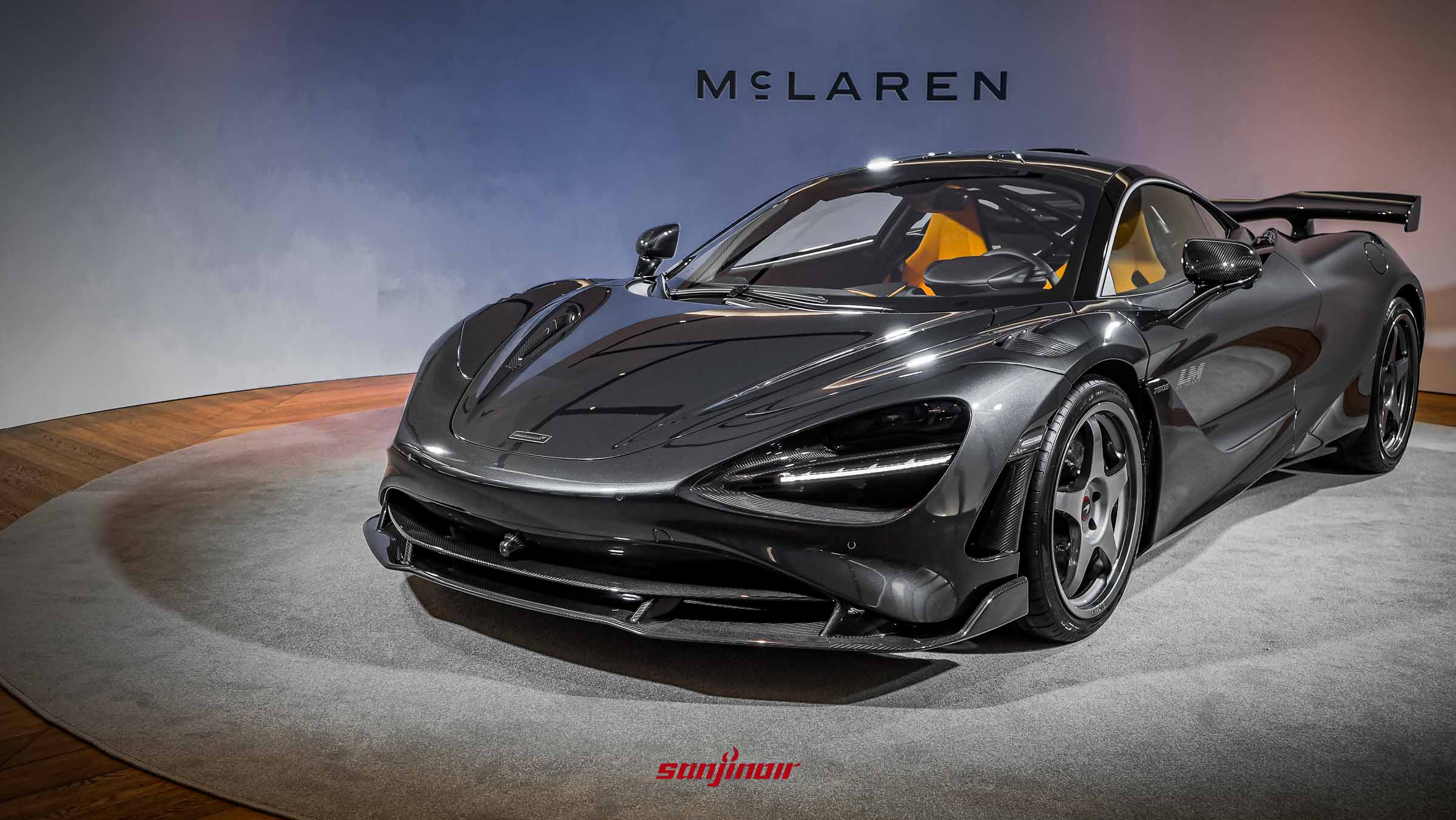 一輛深灰色的McLaren 750S Le Mans跑車停在發表會展示台上，從車頭右前方視角拍攝，展現流線車身與碳纖維前保桿設計。