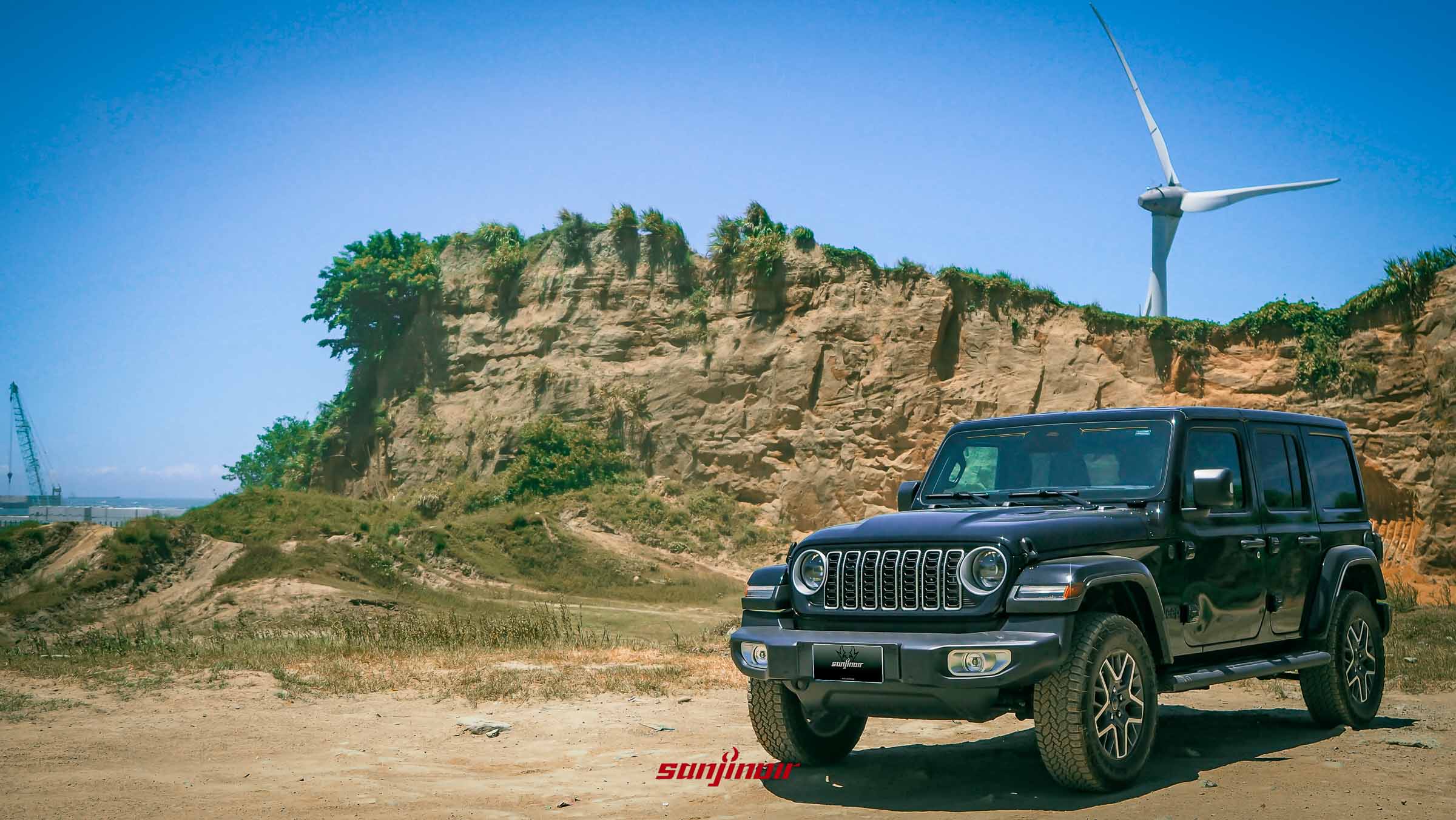 黑色 2026 Jeep Wrangler Sahara 停在黃土坡前，背景有一座風力發電機。