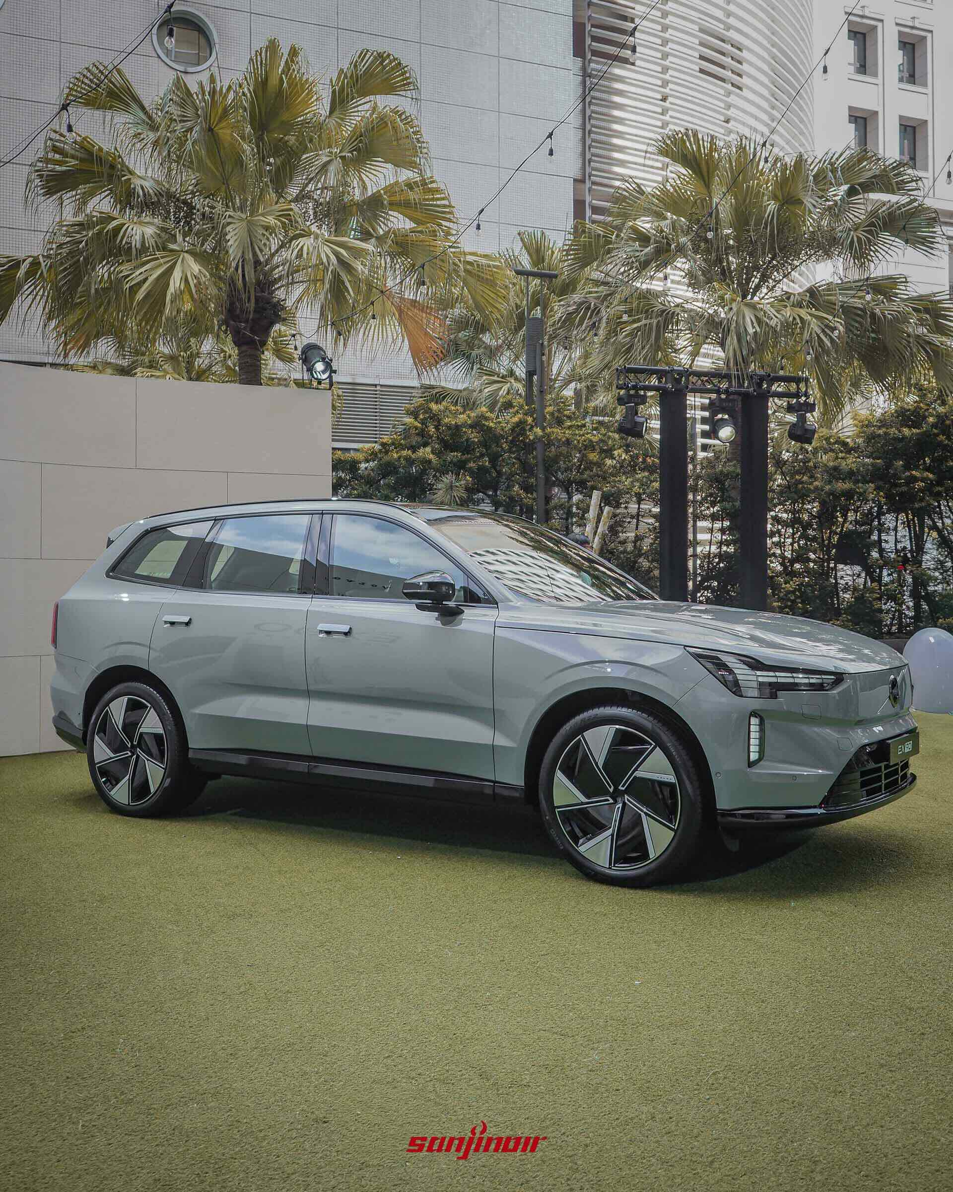 灰色Volvo EX90的車側全景,展示其平滑的車身線條、隱藏式車門把手以及獨特的雙色空氣力學輪圈,背景有棕櫚樹與建築物。