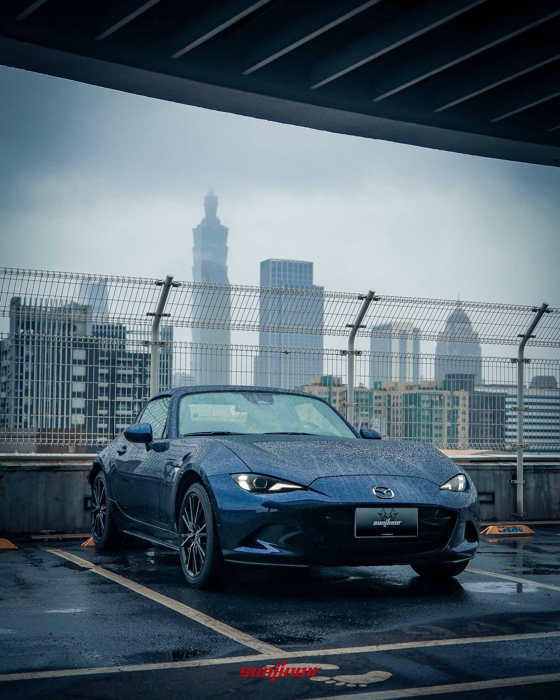 Mazda MX-5 RF 車頭側視圖,車身佈滿雨滴,背景為都市高樓建築與陰雨天空。