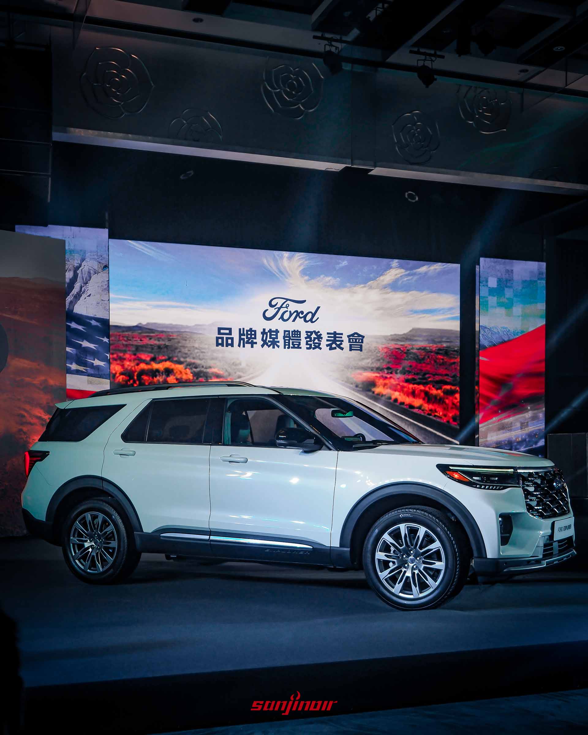 發表會舞台上一輛白色Ford Explorer的側前45度角近景，車身光潔，細節清晰。背景是顯示「Ford 品牌媒體發表會」和美國國旗圖案的大螢幕。