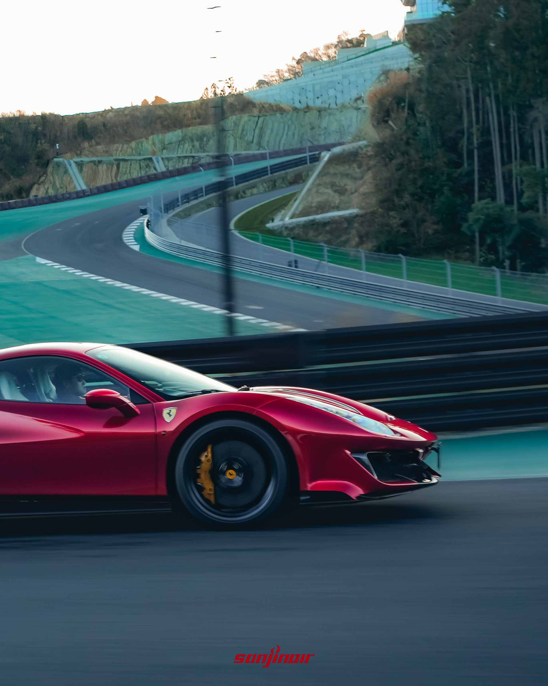 紅色 Ferrari 488 Pista 行駛於帶有綠色緩衝區的賽道上，背景為依山而建的起伏地形與樹林。