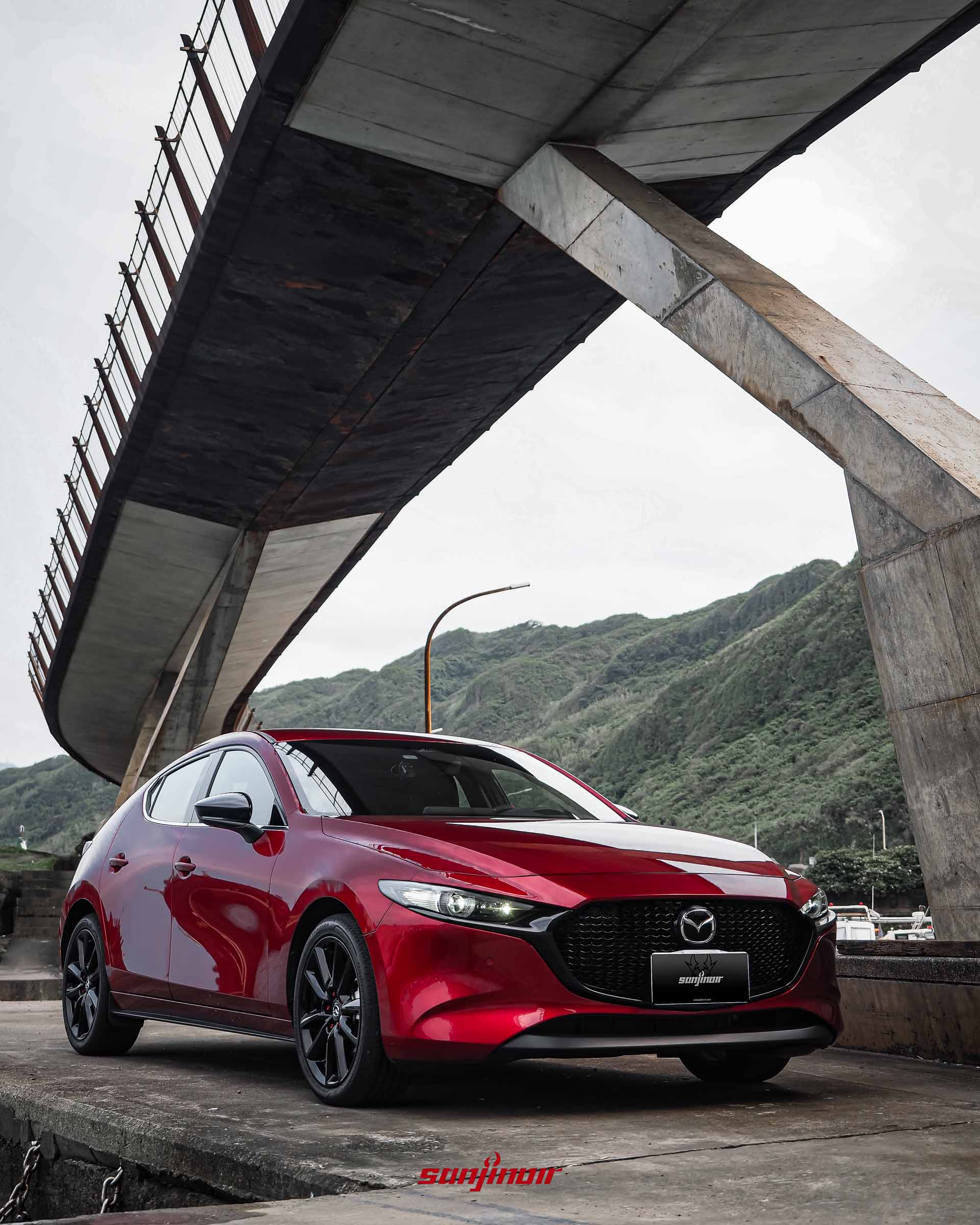 一輛晶艷魂動紅MAZDA3掀背車前部特寫，曜黑水箱護罩與LED頭燈組清晰，停在海邊碼頭。