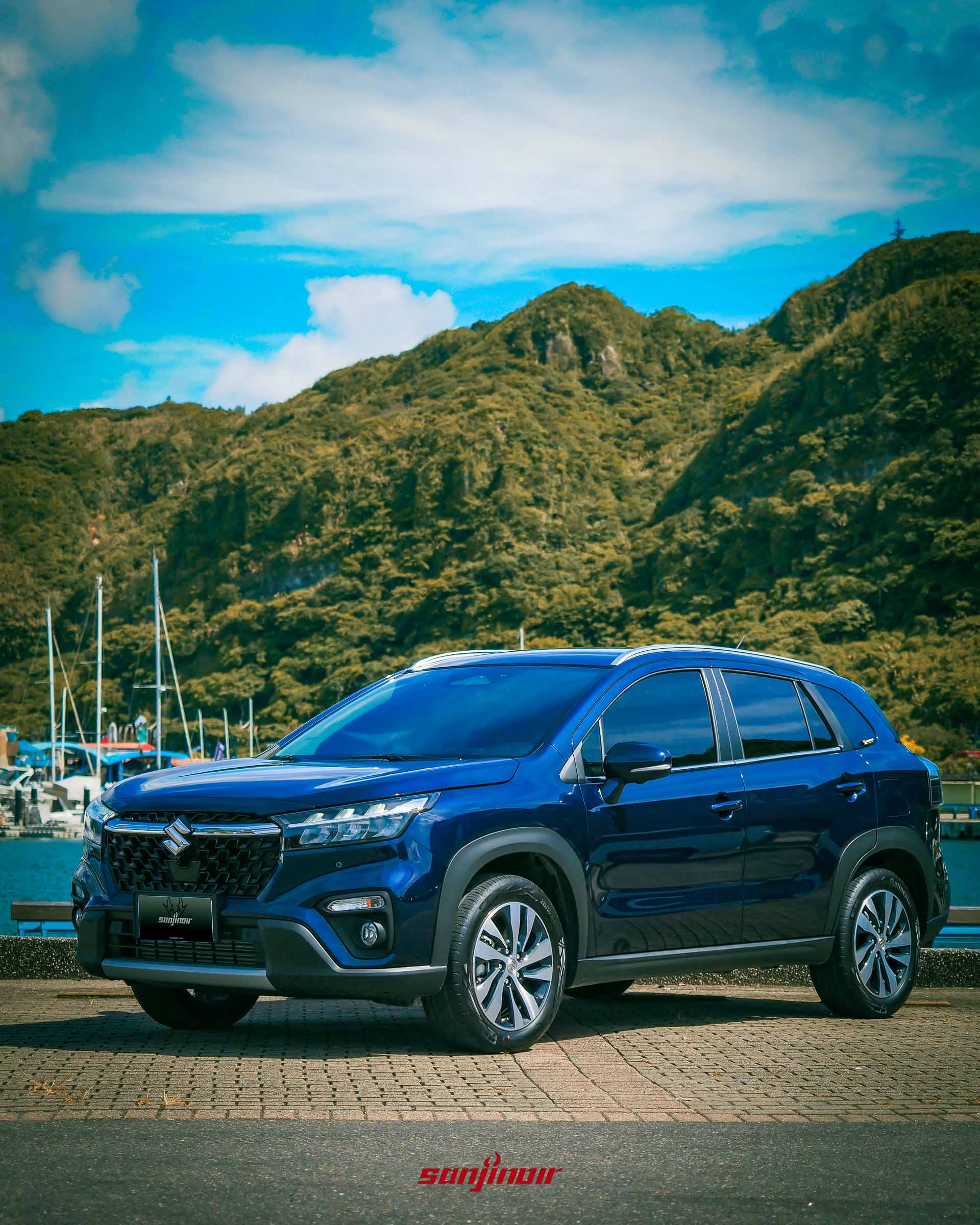 SUZUKI S-Cross Allgrip側前方視角，背景為藍天白雲與綠色山脈。
