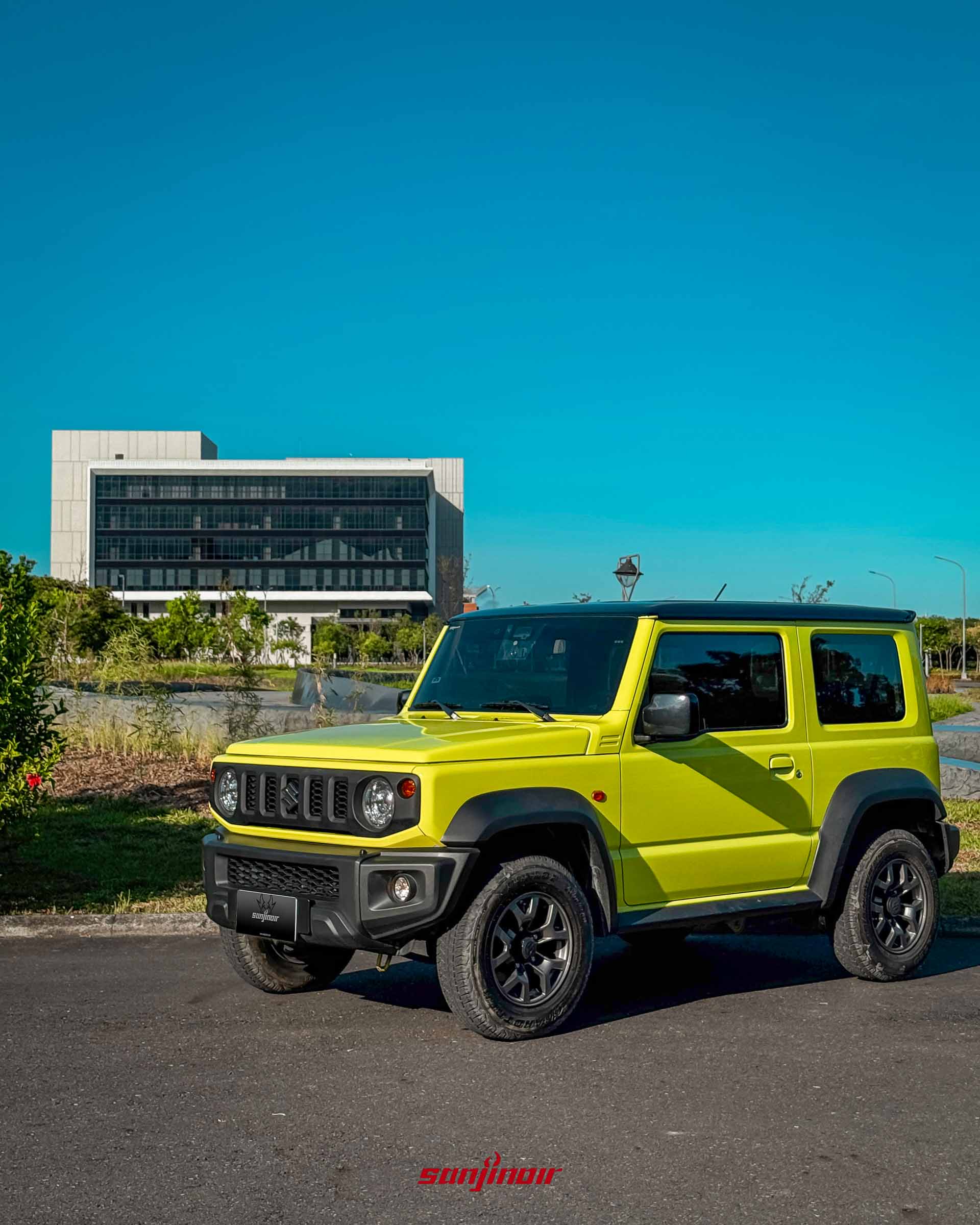 亮檸檬綠 SUZUKI Jimny JB74（配黑頂）的戶外前右側照片，停在柏油路上。背景是一棟擁有大片玻璃窗的現代白色辦公大樓，藍天白雲下有大片綠地。