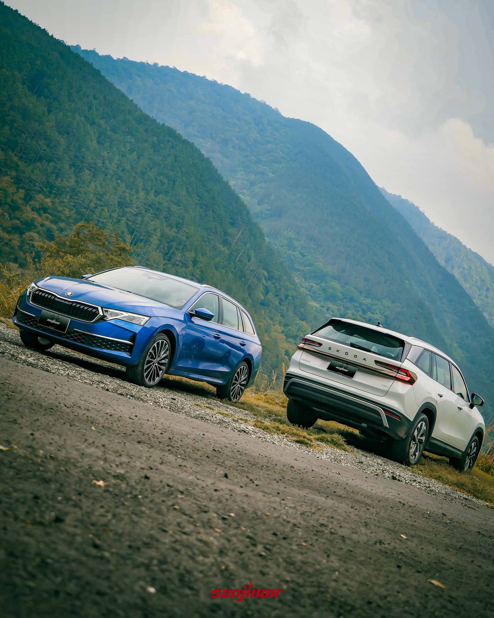 Škoda Octavia Combi 2.0 TSI 4×4（藍色）和 Škoda Kodiaq 2.0 TSI 4×4（白色）並排停在礫石路面上的低視角全景照片，背景是茂密的松樹林和陡峭的山脈。Kodiaq 顯示車尾。