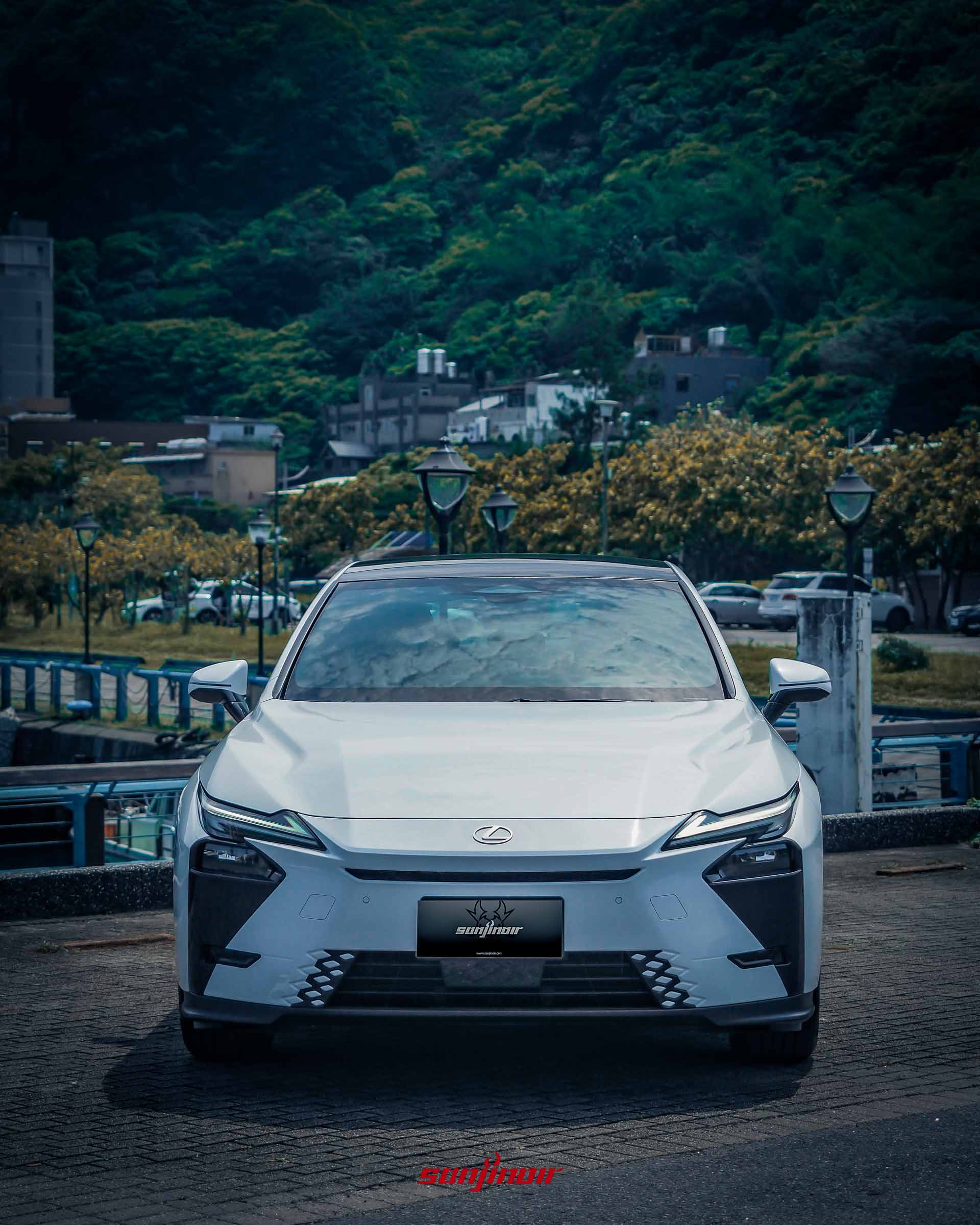 白色Lexus ES 300h的正車頭特寫,展示紡錘型水箱護罩設計與箭矢型LED日行燈,背景為碼頭與山坡。