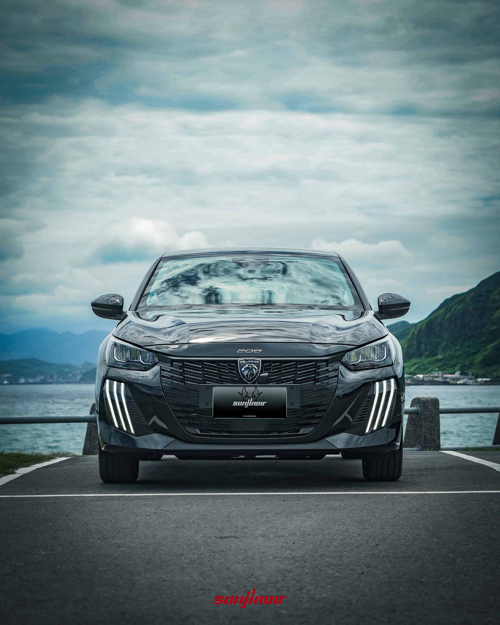 黑色2026年式PEUGEOT 208 Hybrid車頭正視圖搭配海景與多雲天空背景。