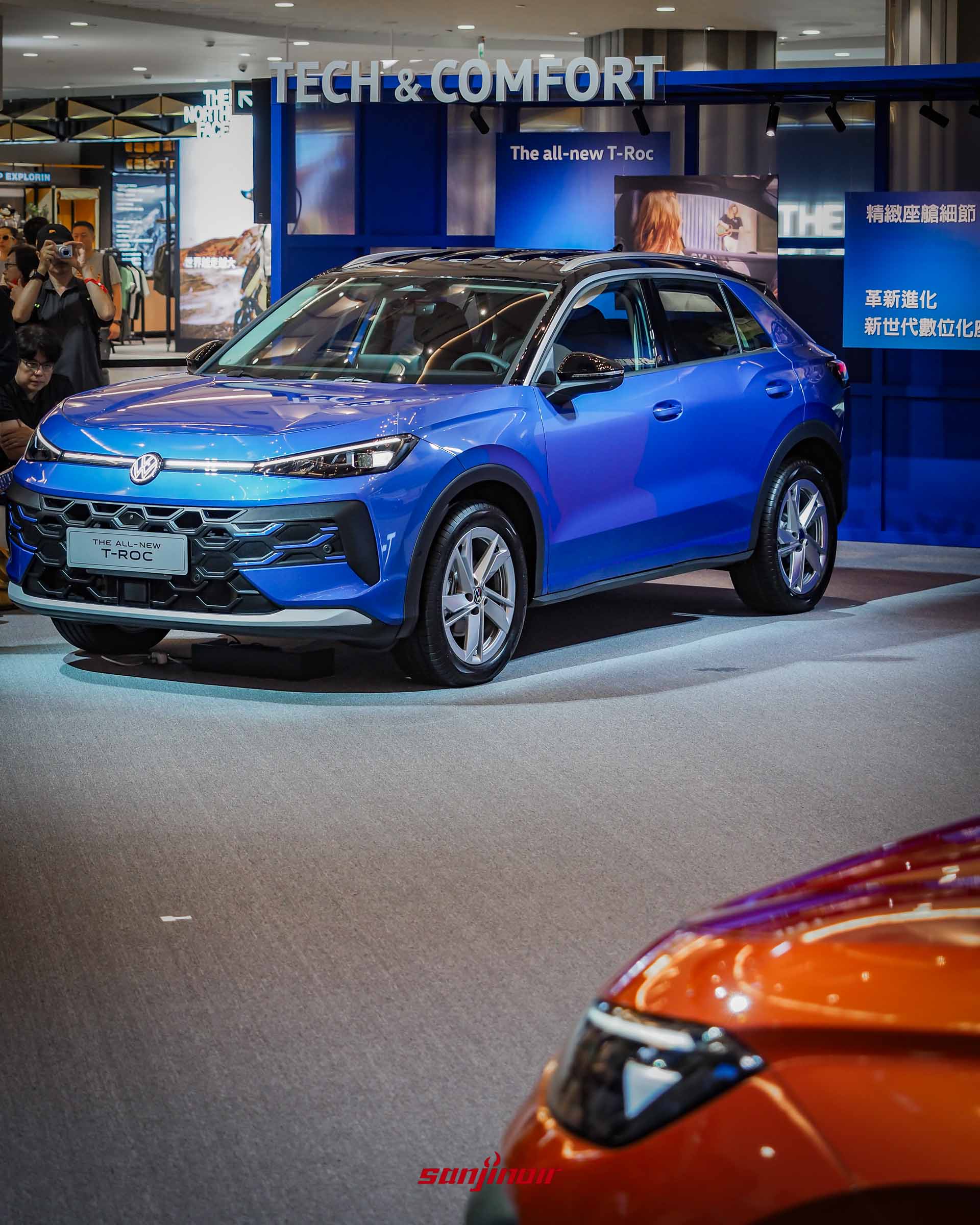 藍色 Volkswagen T-Roc 車頭特寫,清楚展現全新 IQ.LIGHT 矩陣式頭燈與水箱護罩上的貫穿式 LED 定位燈,前景邊緣有另一輛橘色車款的局部。