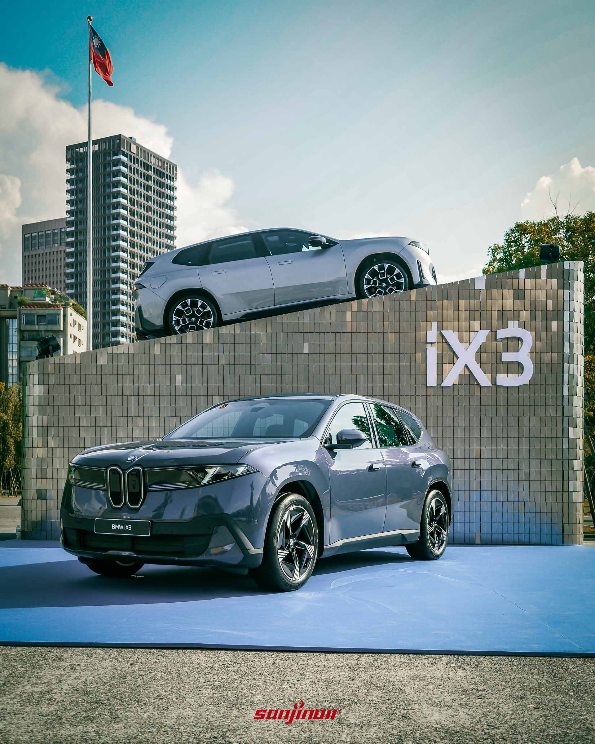 垂直構圖的BMW iX3戶外展區，深藍色車款在前方，銀色車款在上方展台，背景有高樓與一面國旗。