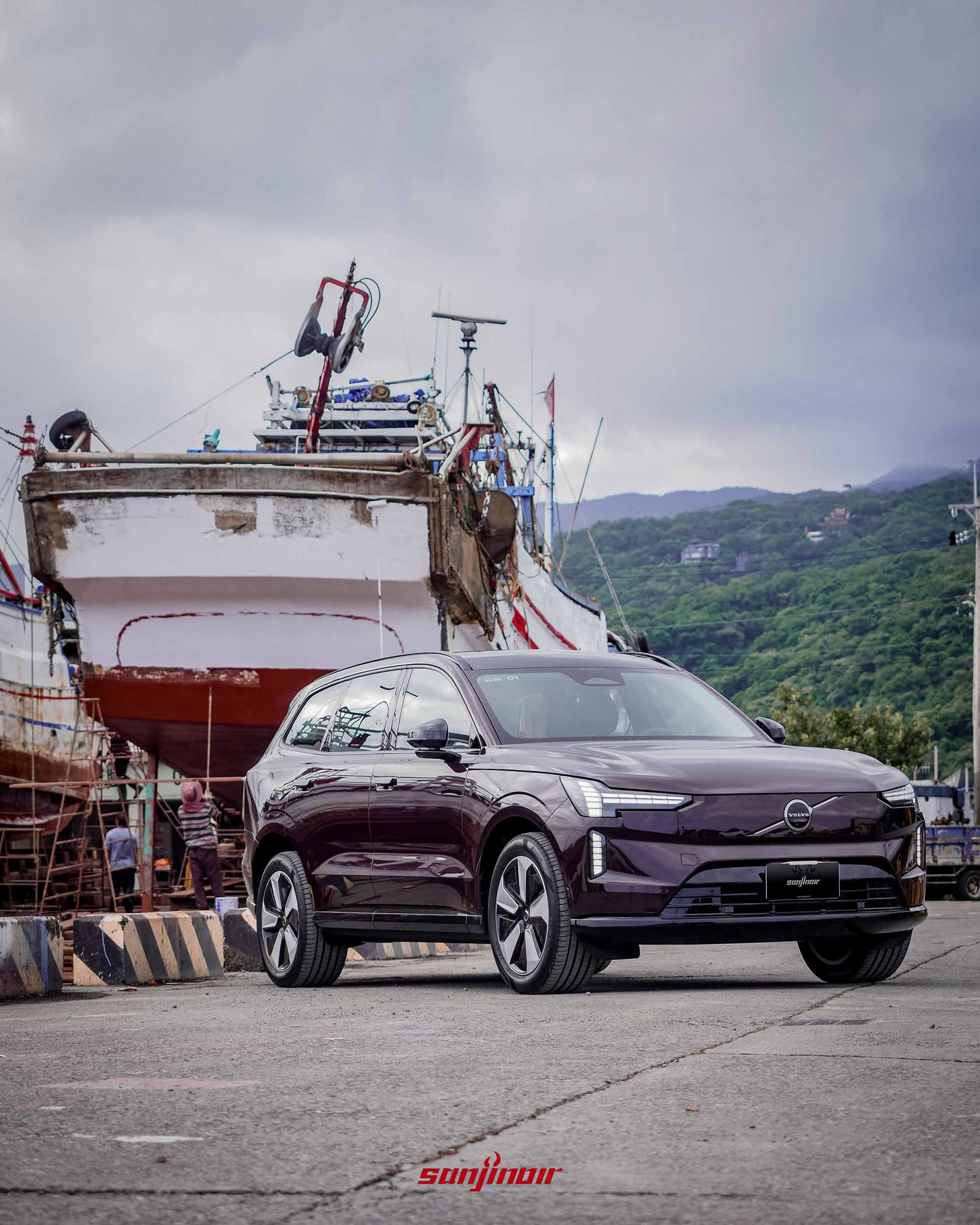 野莓紅Volvo EX90的右前側車身全景照,停放在漁港碼頭,後方有正在維修的漁船與遠方山景。