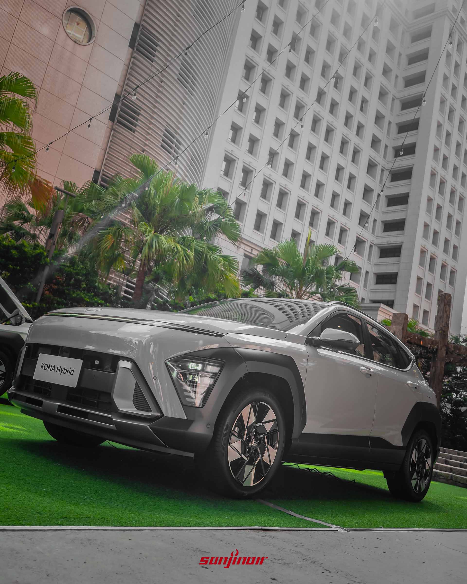 以低角度拍攝淺灰色Kona Hybrid的前側車身。車輛停放於草皮上,配備幾何造型的雙色切削鋁圈與大面積黑色防刮輪拱,背景為高聳的現代白色建築物與熱帶棕櫚樹。