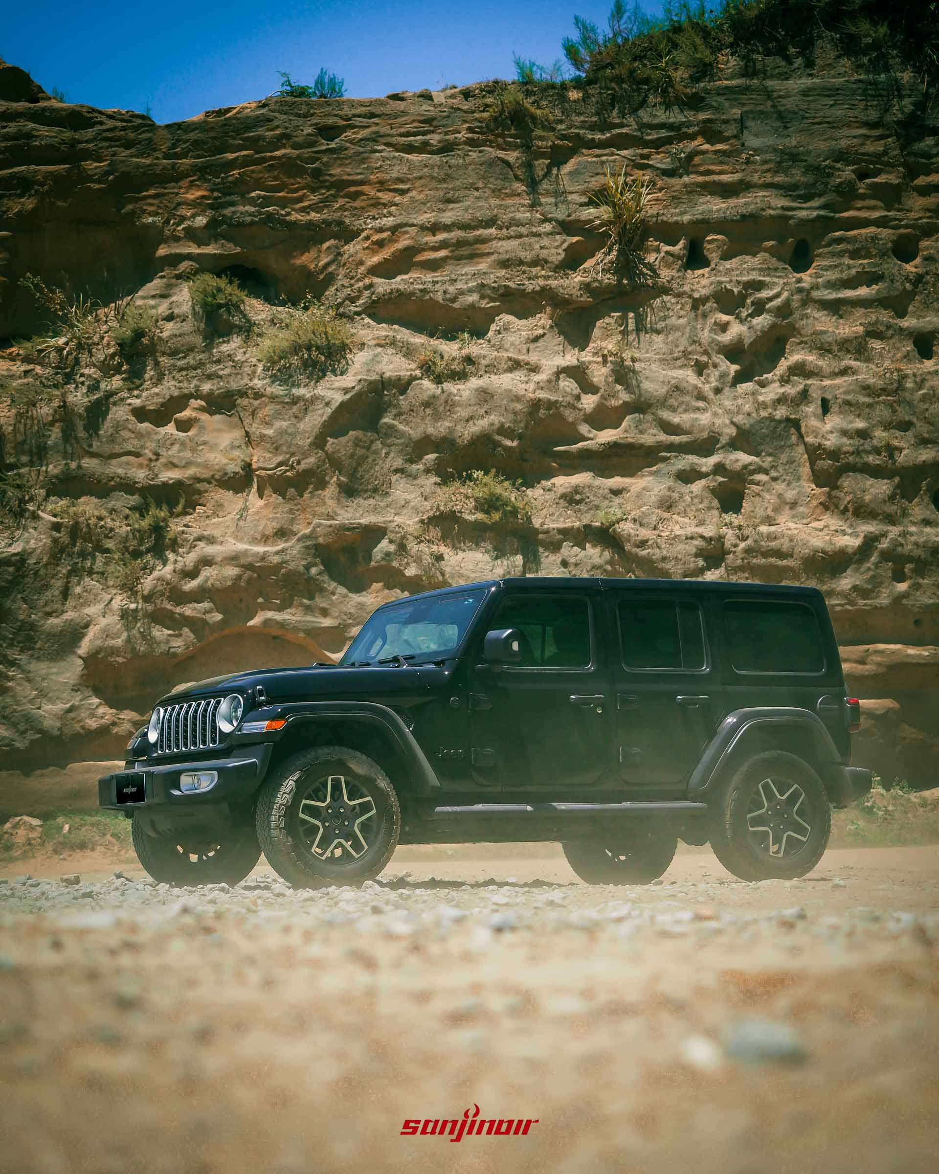 黑色 Jeep Wrangler Sahara 的側面照,停在陡峭的岩壁前,展現粗獷的車身輪廓。