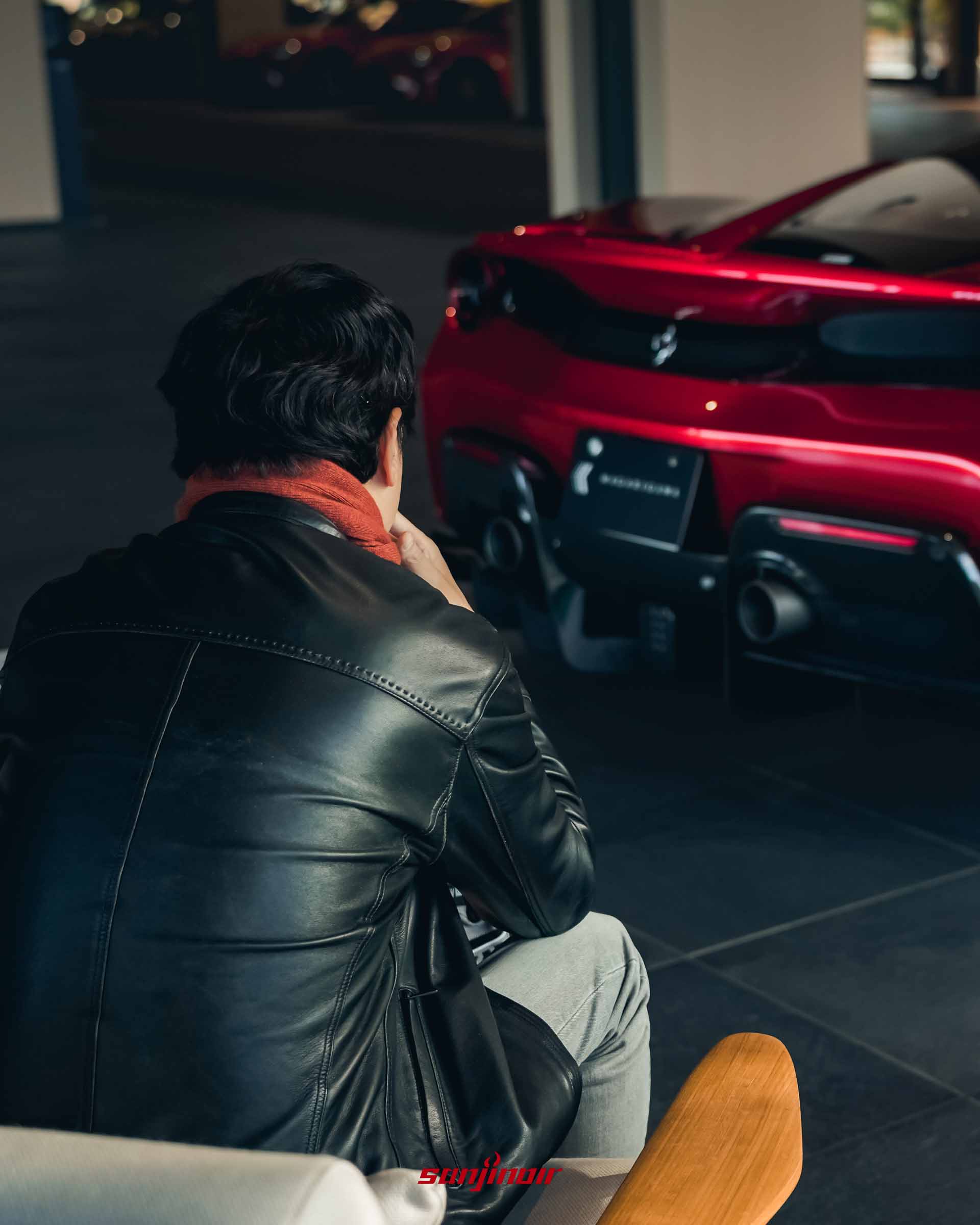 一名男子的背影，正坐在沙發上望向窗外停放的紅色 Ferrari 跑車車尾。