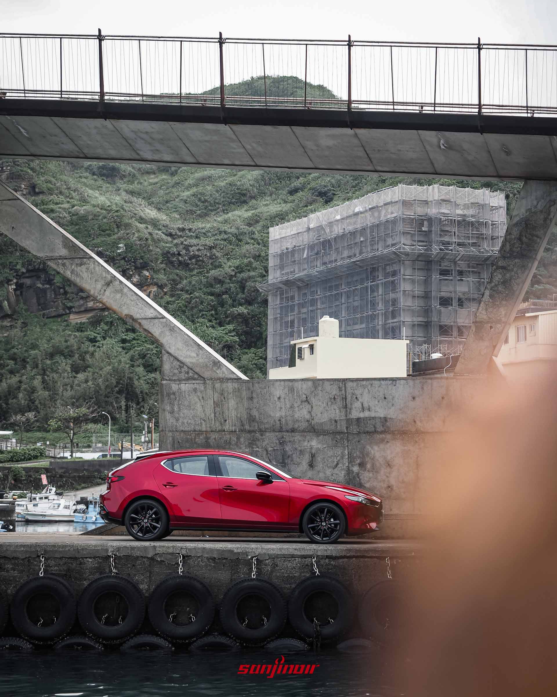 一輛晶艷魂動紅MAZDA3掀背車側面圖，停在海邊碼頭，背景是混凝土高架橋和一座建築物，前景有模糊的遮擋物。
