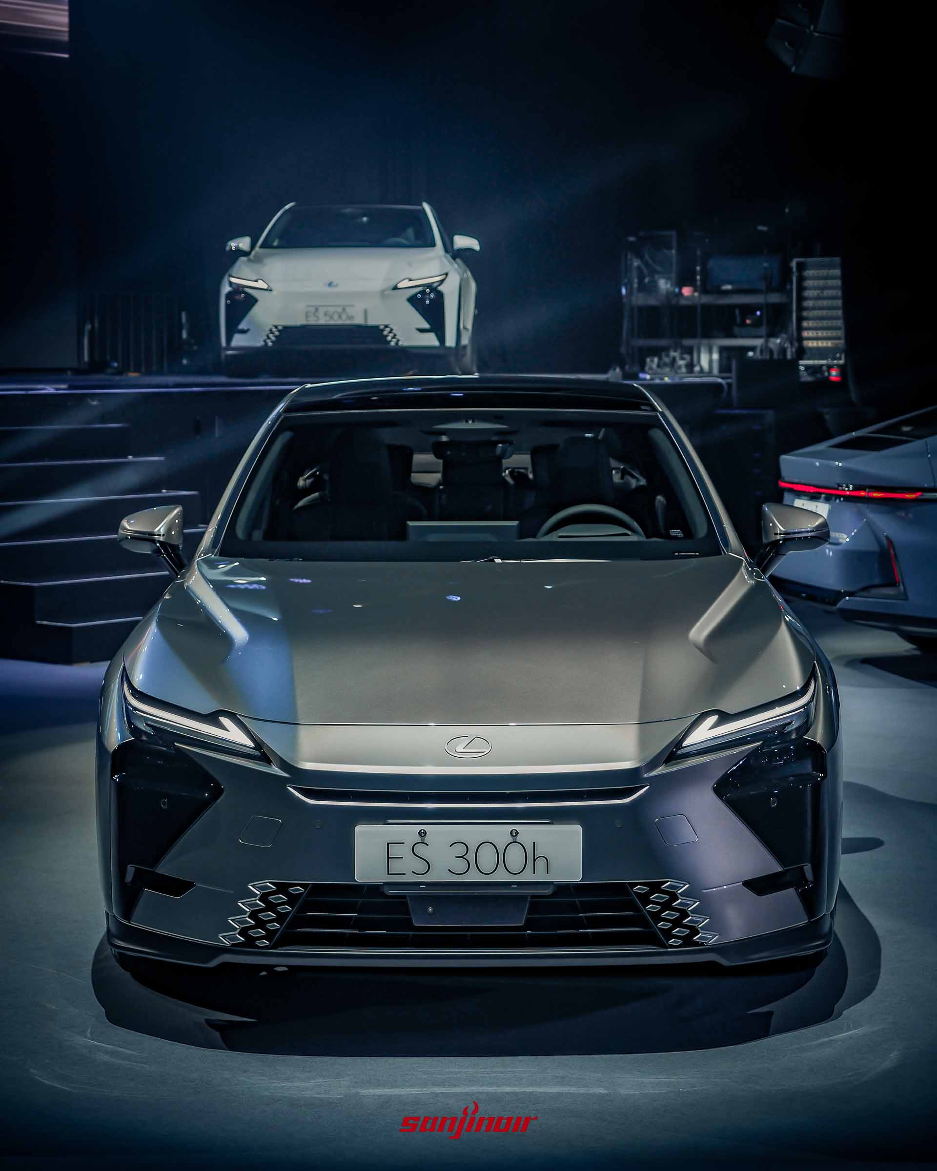 銀色Lexus ES300h正面特寫，展示紡錘型水箱護罩設計與LED頭燈組，背景有一輛白色車款。