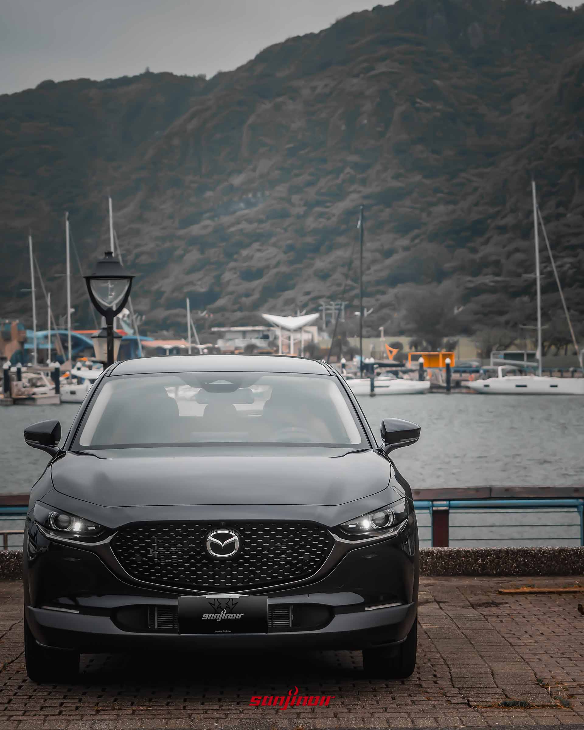 黑色MAZDA CX-30 20S Homura車頭正視圖,開啟LED頭燈,背景為遊艇碼頭與翠綠山巒。
