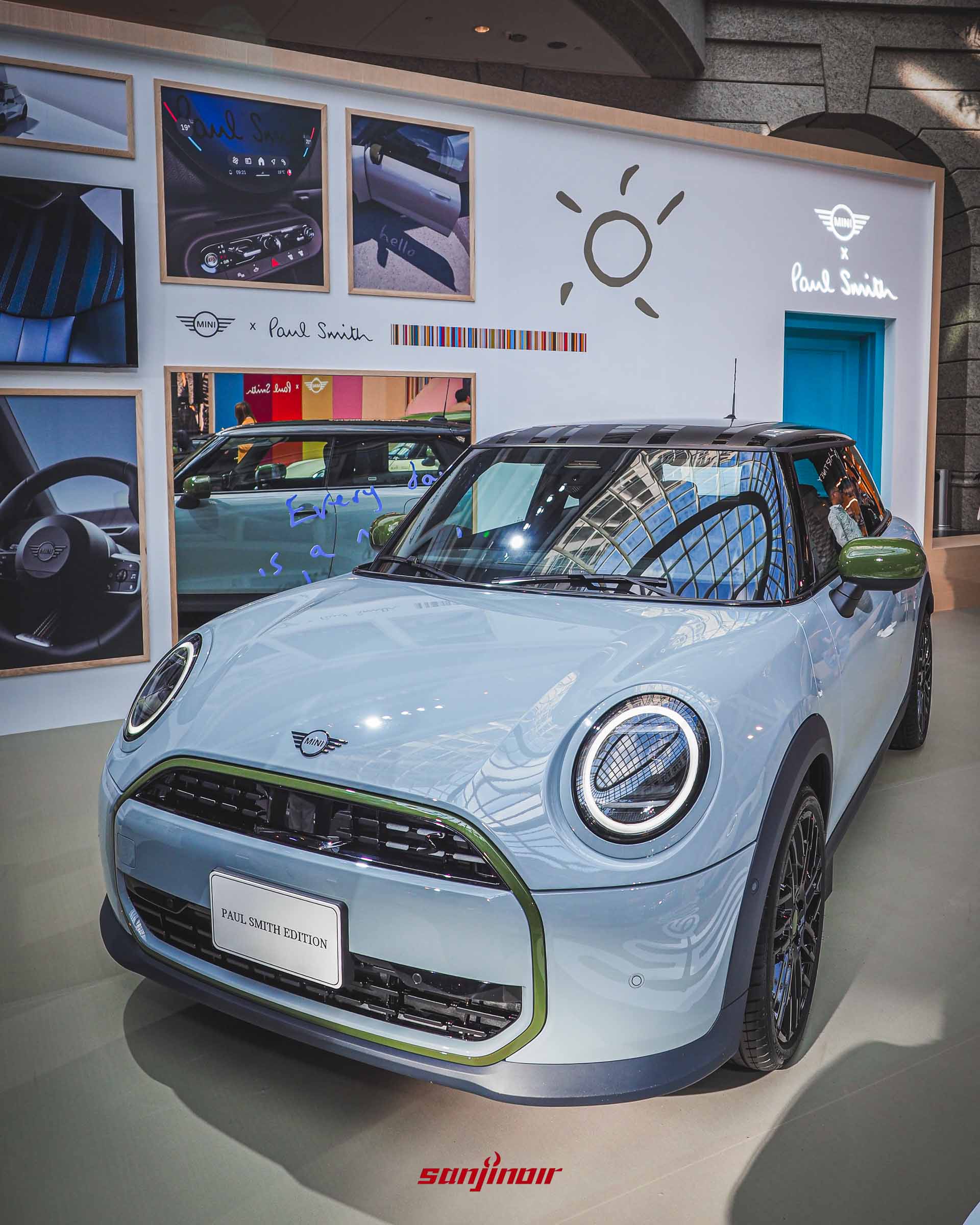 一輛灰藍色塗裝的MINI特仕車停在展示區，背景是一面白色展示牆，牆上有太陽塗鴉、手寫字跡與數張內裝和車體細節的相框。