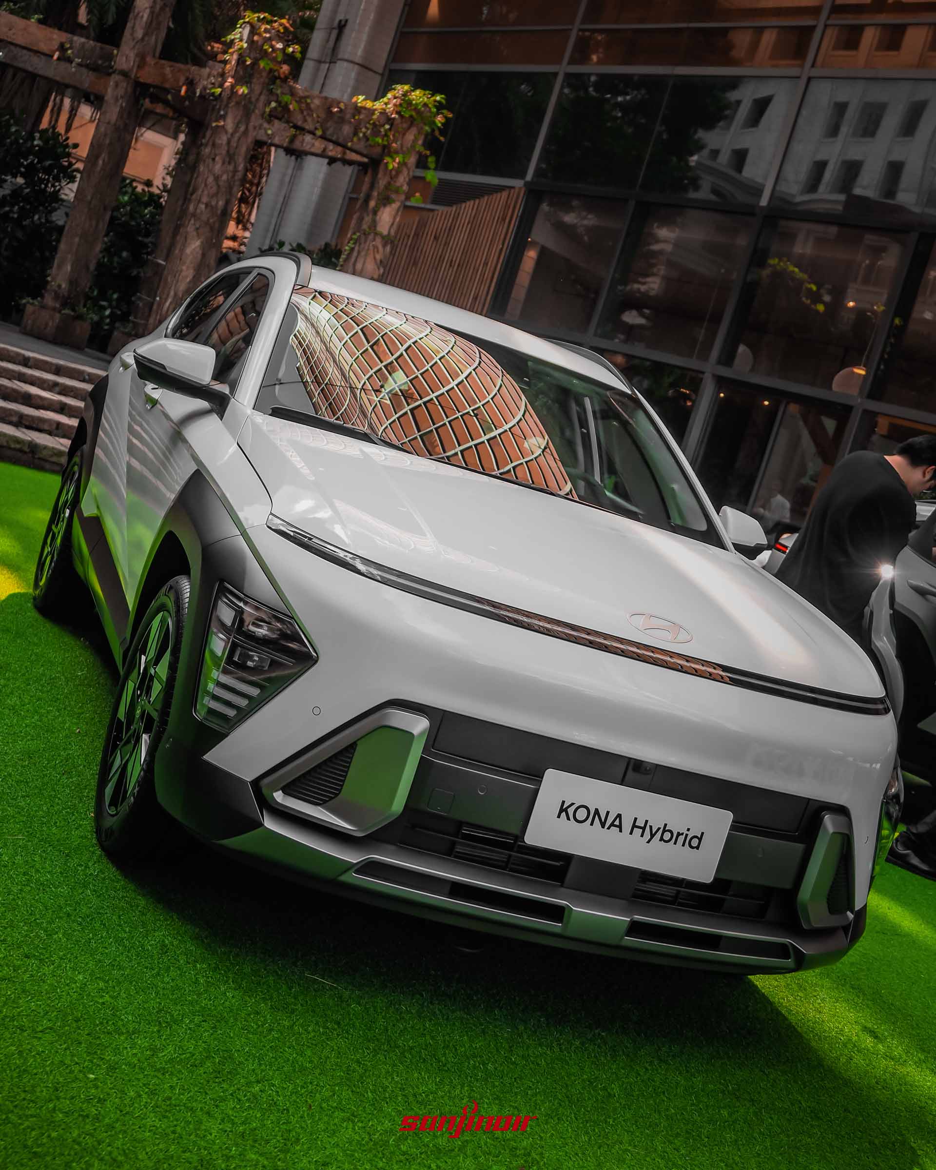 近距離俯拍Kona Hybrid的車頭細節。畫面聚焦於貫穿車頭的Seamless Horizon Lamp無縫地平線LED日行燈、隱蔽式頭燈組以及下方的幾何進氣壩,車牌位置懸掛「KONA Hybrid」銘牌。