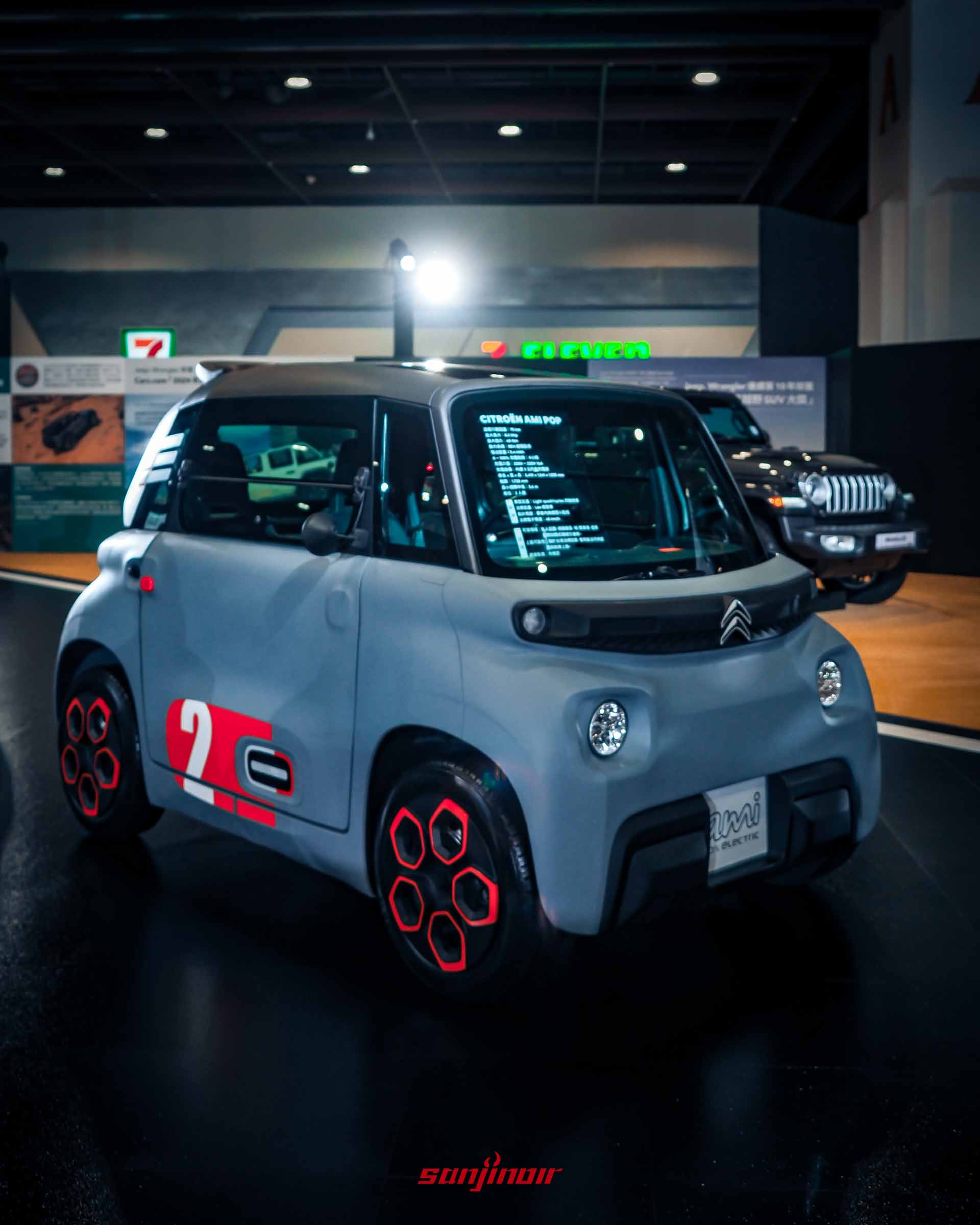 灰藍色Citroën Ami微型電動車，車門上有紅色與白色數字塗裝，背景為Jeep Wrangler。