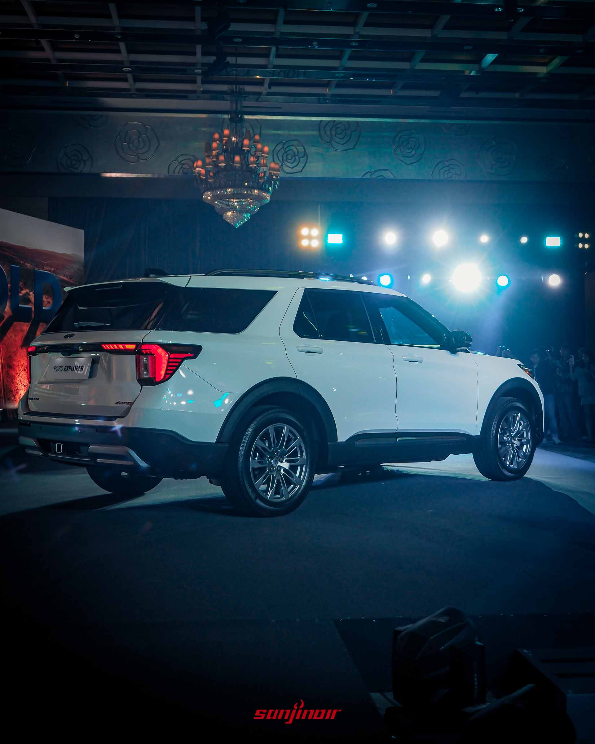 發表會舞台上一輛白色Ford Explorer的後45度角全景。車尾有「FORD EXPLORER」車牌和「AWD」標誌。背景大螢幕顯示「BOLD」沙漠圖案，上方有一個華麗的水晶吊燈和舞台燈光。