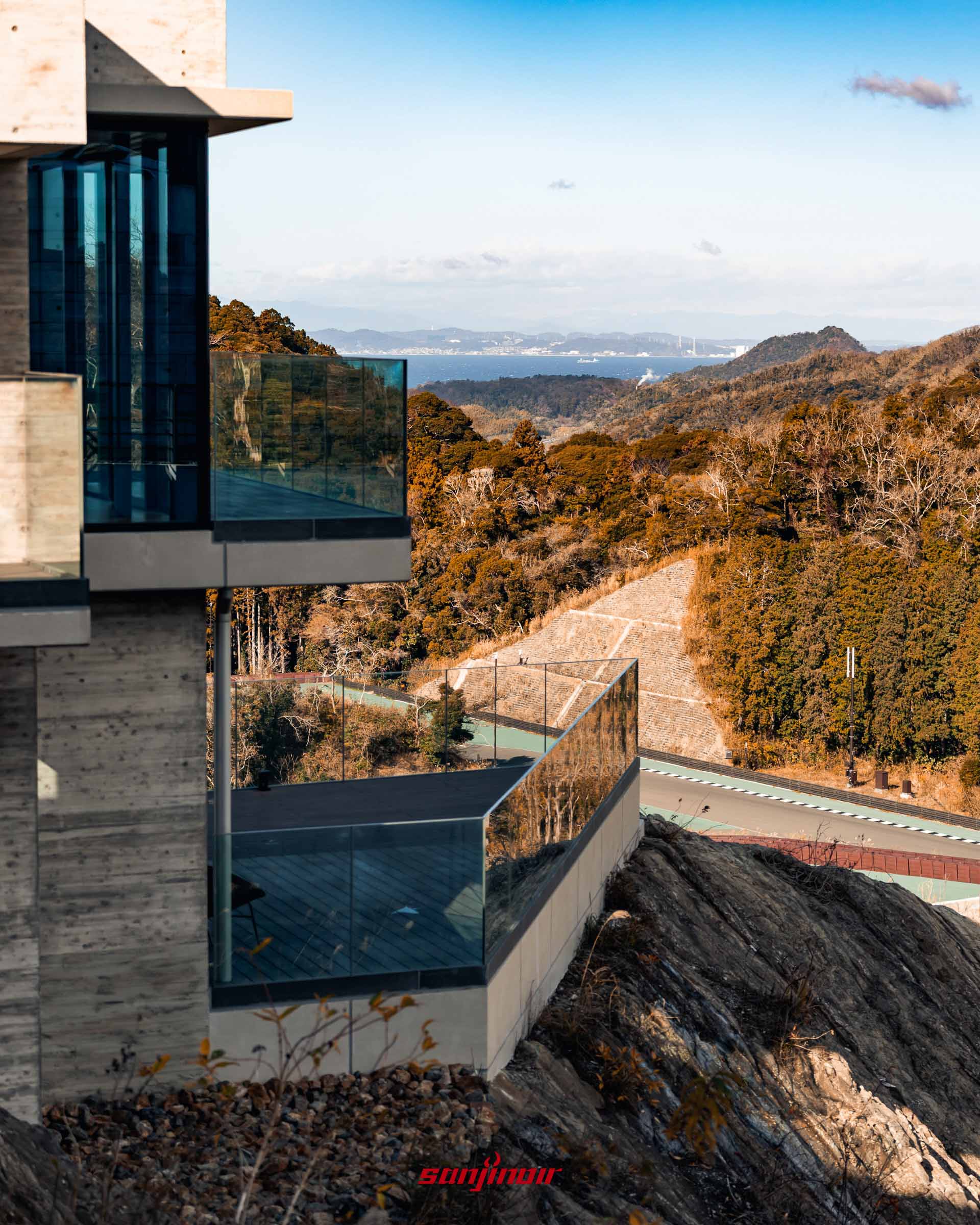 現代風格的別墅建築外觀，擁有大面積玻璃陽台，建於岩石山坡上，可俯瞰下方賽道與遠處的海灣。