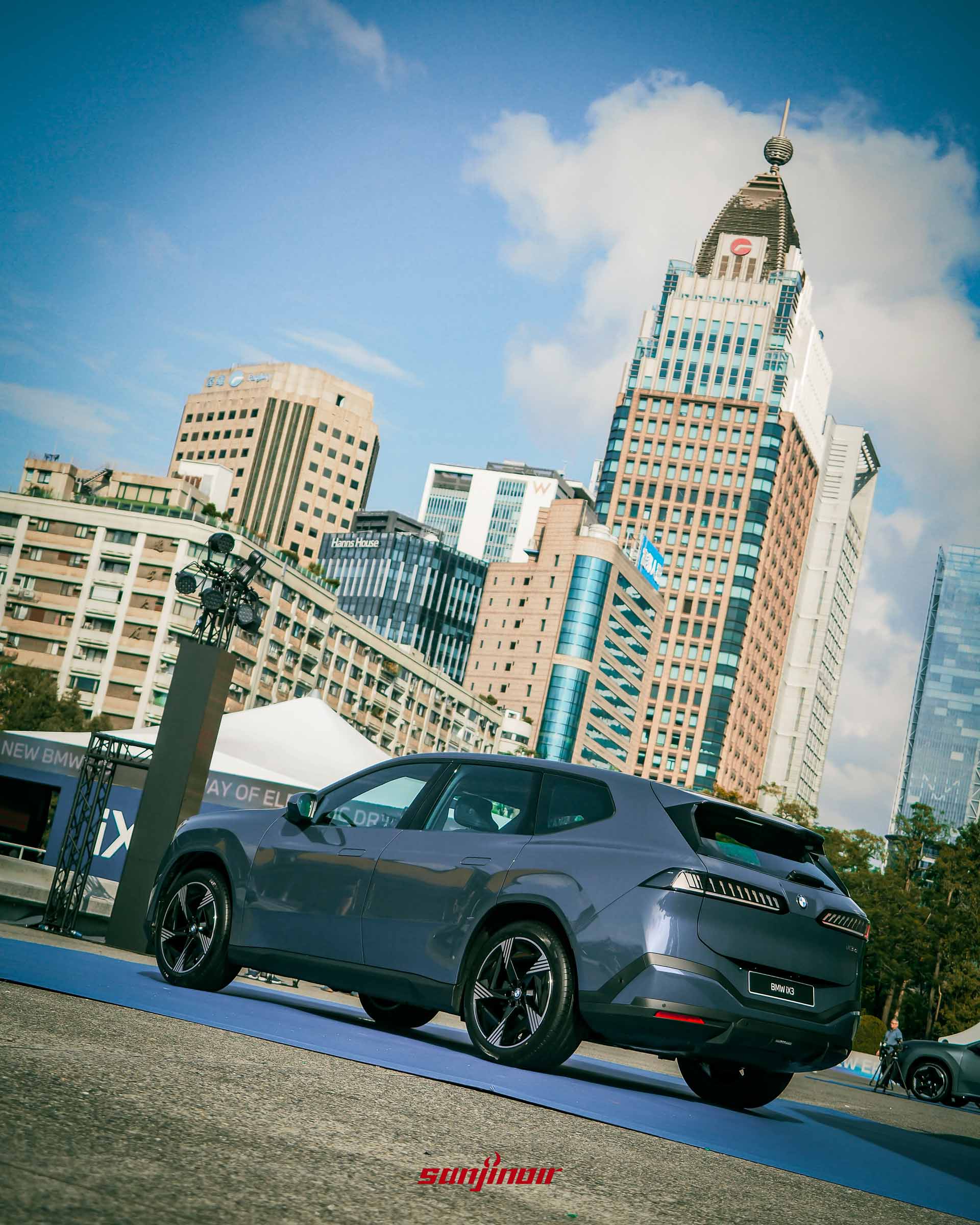 Olympus Digital Camera深藍色BMW iX3右後方視角，展示流線車身與獨特斜向尾燈設計，背景為台北都會高樓群。
