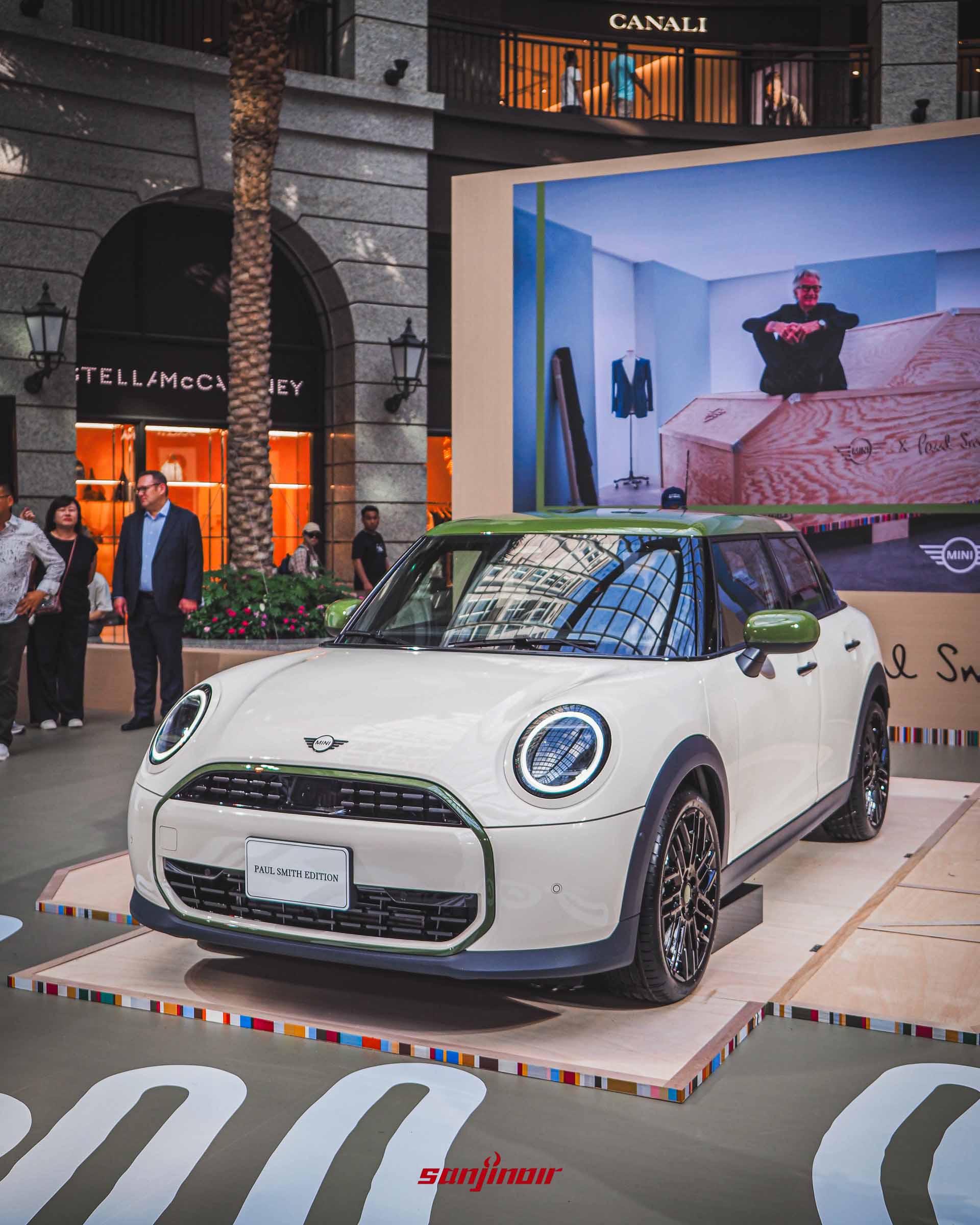 白色MINI特仕車停在商場走道中央的展示台上，背景設置了一面大型輸出牆，畫面上是設計師Paul Smith坐在印有聯名標誌木箱上的照片。