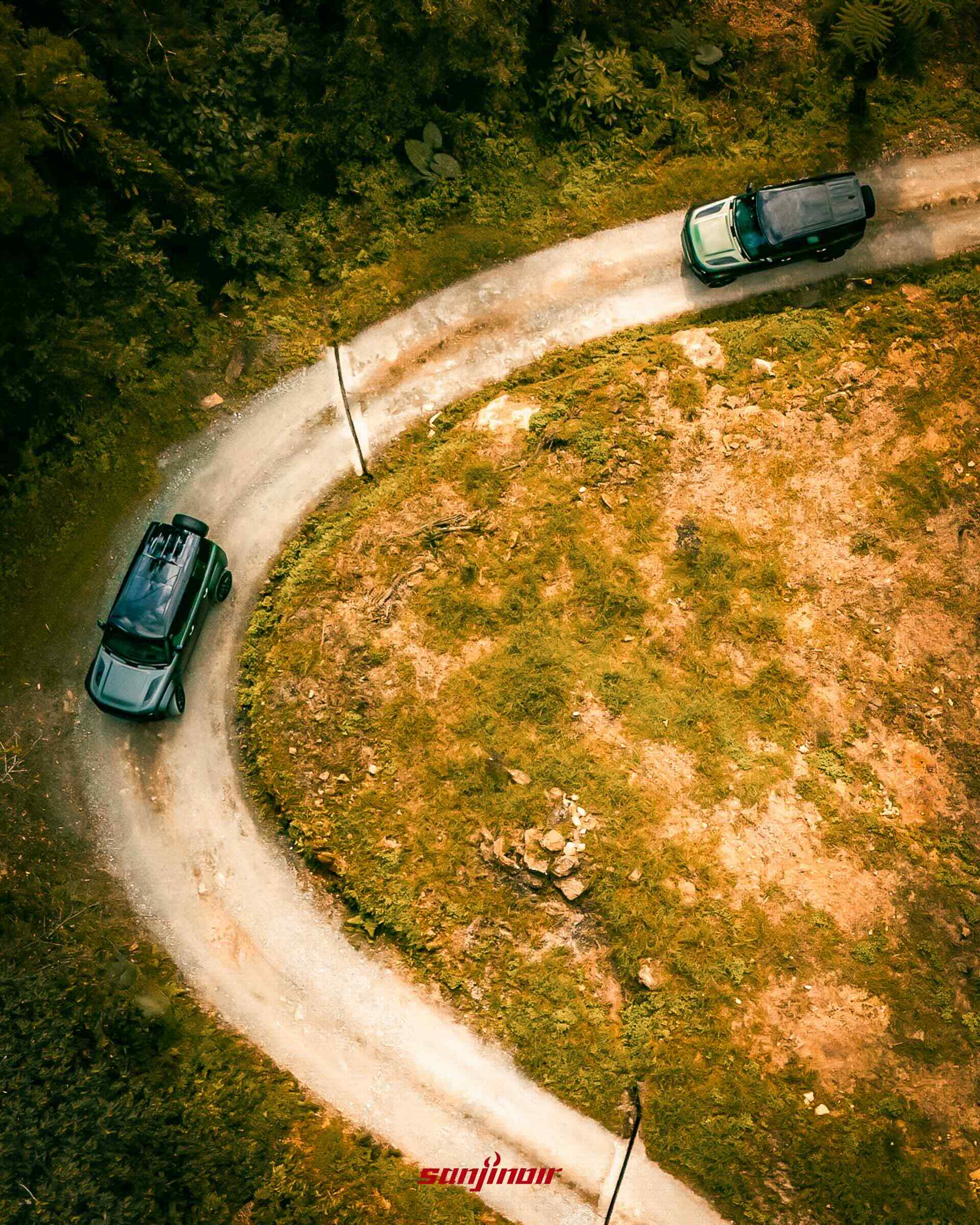 俯瞰視角拍攝兩輛Defender行駛於蜿蜒林間碎石路上。