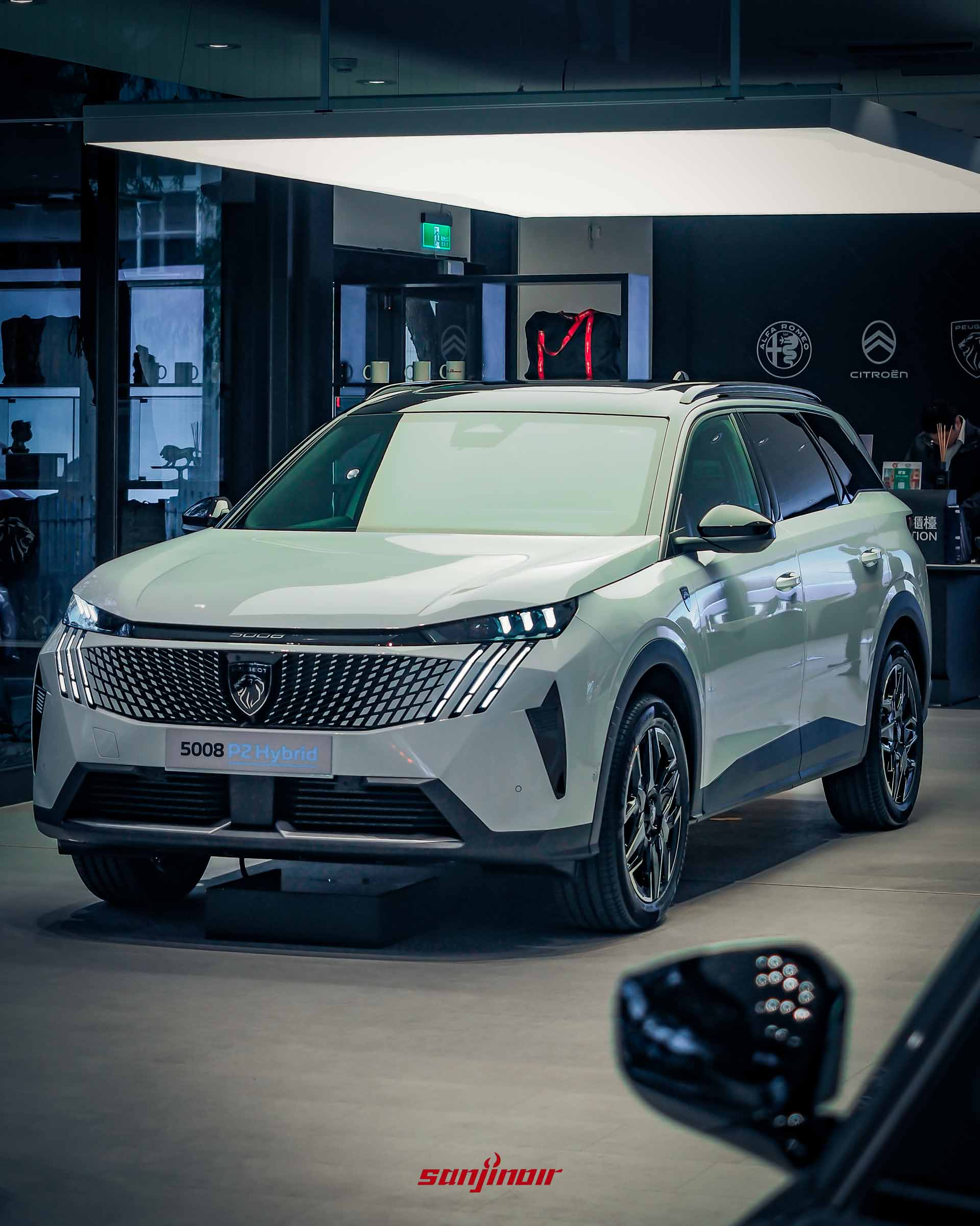 一輛白色的 PEUGEOT 5008 P2 Hybrid SUV。位於發光的燈板下方。車輛具有標誌性的爪痕 LED 大燈，和巨大的車頭格柵。