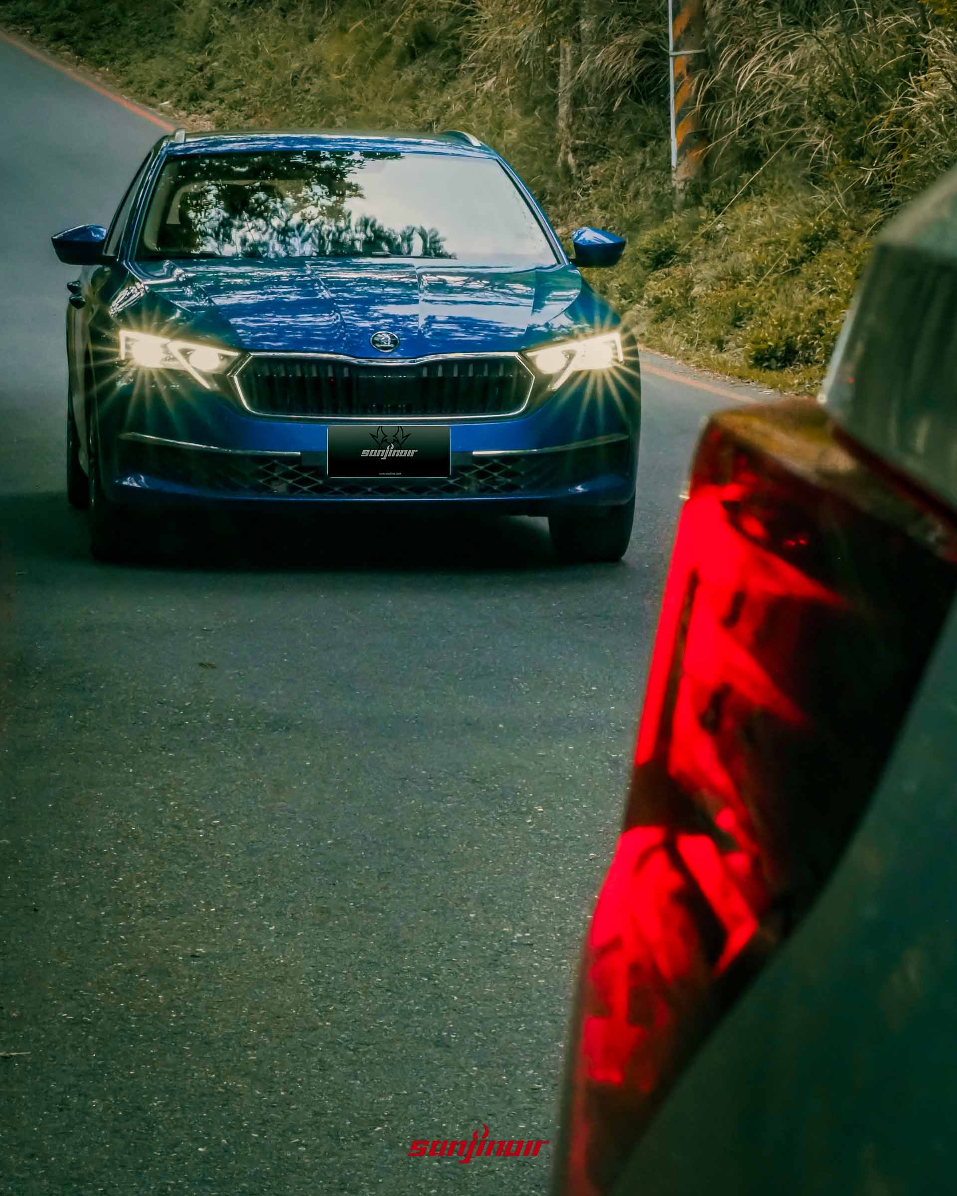 藍色 Škoda Octavia Combi 2.0 TSI 4×4 行駛在山路上的正面照片，右側前景有模糊的紅色尾燈作為視覺遮擋物。