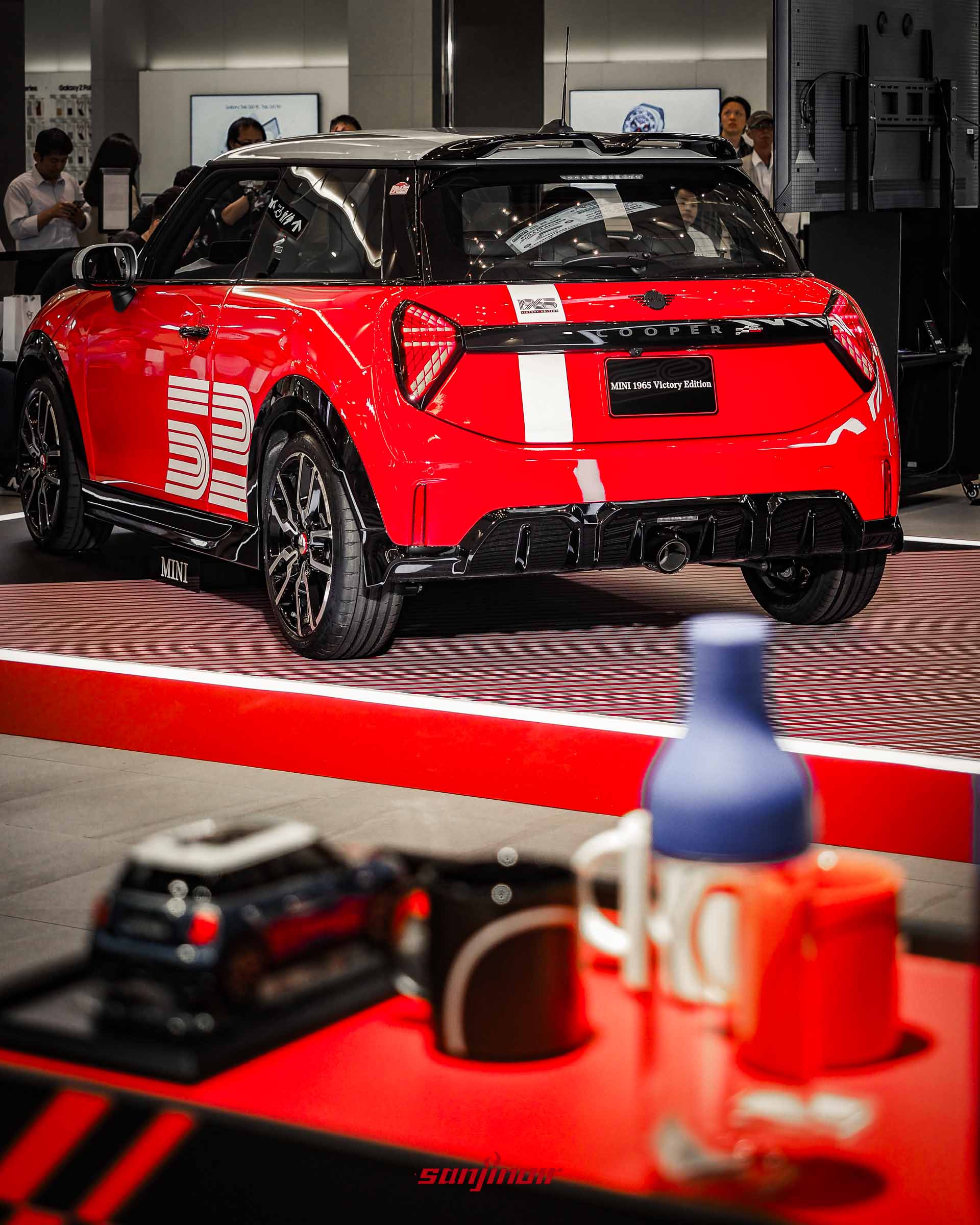 MINI 1965 Victory Edition的右後方視角,展示車尾的白色飾條、尾燈以及下方的黑色擾流板,前景有周邊紀念商品。