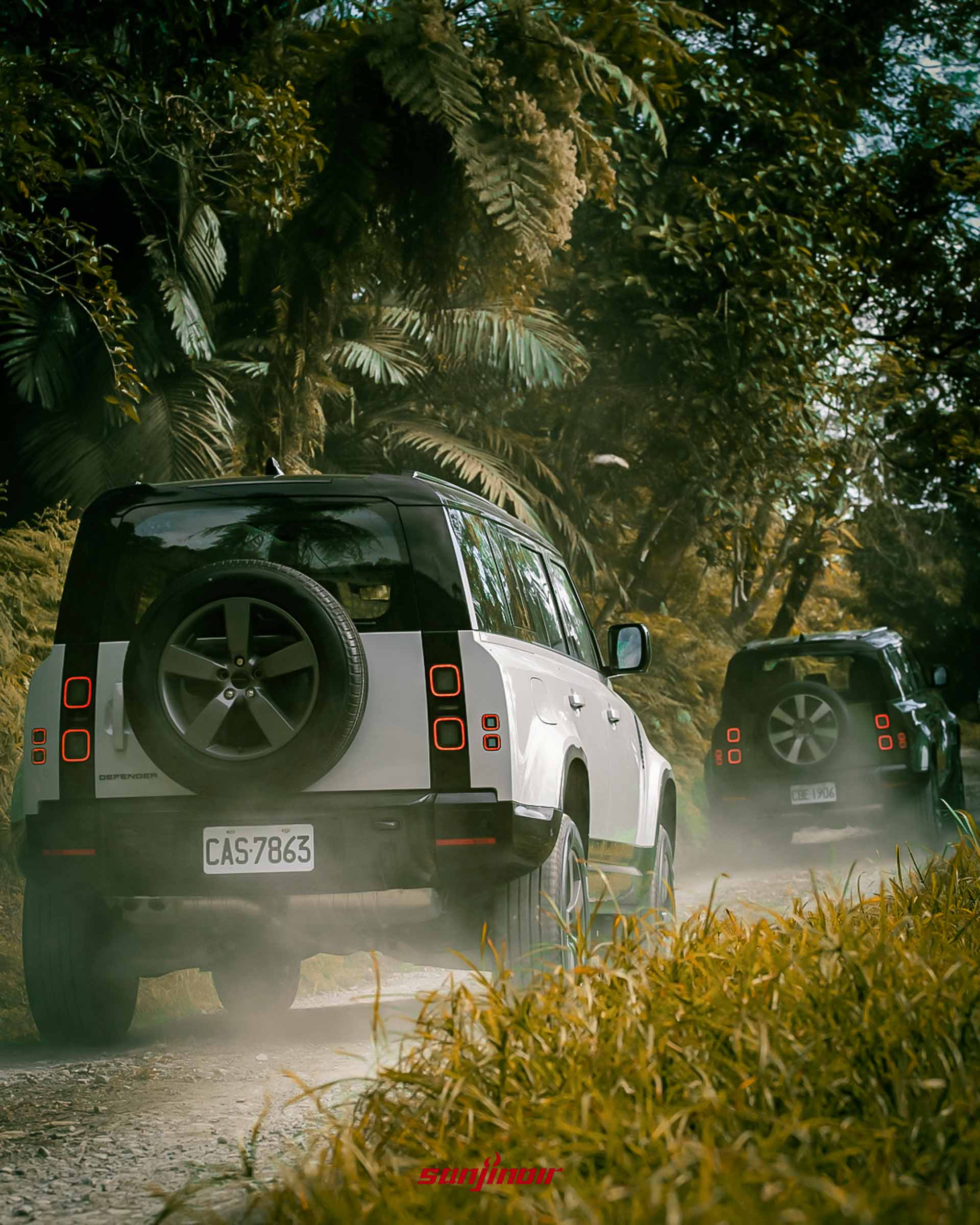 兩輛Defender行駛於泥土路上車尾揚起沙塵兩旁為茂密樹林。