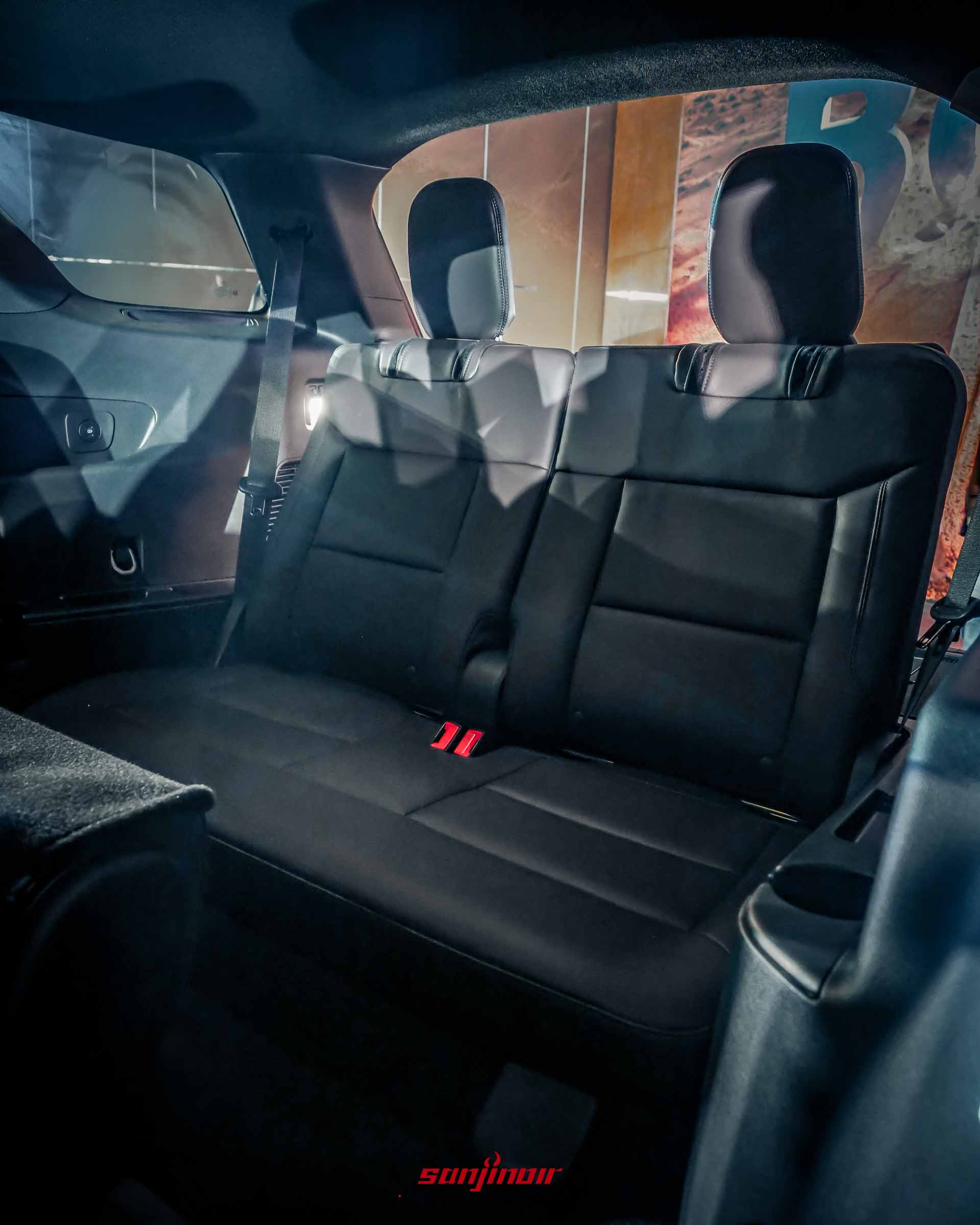 Ford Explorer車內特寫，展示黑色皮革的第三排座椅。紅色安全帶扣環清晰，頭枕可見。右側D柱有空調出風口和杯架細節。背景是大螢幕上的沙漠「BOLD」字樣。